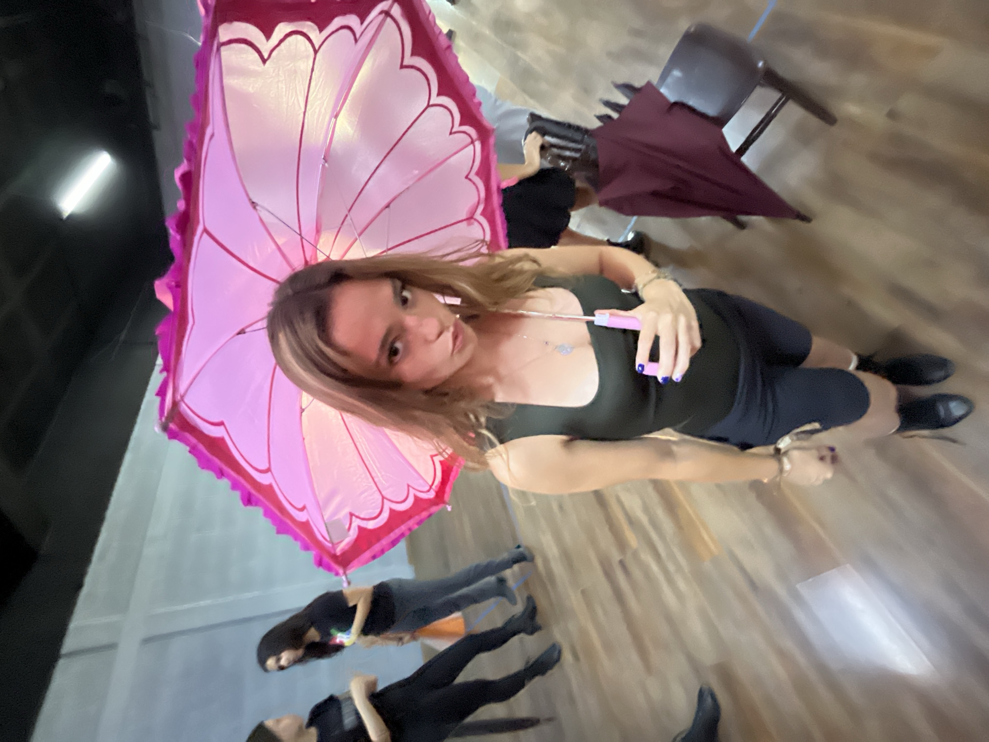 The image is an angled, high-vantage point shot of a woman holding a large, pink, flower-shaped parasol. She is wearing a dark-colored, low-cut top, dark bottoms, and black shoes, and she has fair skin and brown hair. She is holding a pink object in her hand with purple nail polish.
In the background, there are several other people lying on the floor, some wearing black and others wearing gray. There is also a dark-colored chair and a maroon-colored object on the floor. The floor appears to be made of wood.
Given the lack of clear landmarks or identifying features, it is not possible to verify if the image was taken in San Bernardino Tlaxcalancingo, Mexico, based on the provided image.