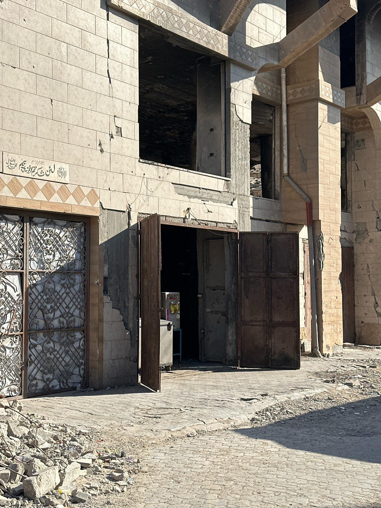 The image shows a close-up, eye-level view of the facade of a damaged building, likely in the city of Gaza, Palestinian Territories, on a sunny day. The building is constructed with light-colored stone tiles, many of which are cracked or missing, revealing underlying brickwork and structural damage. There are also numerous small pockmarks on the facade, possibly from shrapnel.
In the foreground, a cobblestone street is littered with debris, including shattered concrete, rebar, and miscellaneous rubble, indicating recent destruction. To the left, an ornate, wrought-iron gate covers an entrance, with intricate scrollwork patterns. Adjacent to it, a dark metal door stands open, revealing a glimpse of the interior, which appears to be a storage area with shelves and some equipment, including a refrigerator-like unit. To the right of this open door, a large, heavy, dark metal double door is also open, its surface weathered and worn.
Above the entrances, a band of decorative tiles featuring a diamond pattern runs horizontally. Higher up on the building, large window openings are visible, some completely devoid of glass and showing dark, empty spaces within. Architectural details, such as arched openings and stone columns with decorative friezes, are visible on the upper floors, suggesting a building of some architectural significance, possibly a religious or governmental structure. The overall impression is one of severe damage and neglect, consistent with the aftermath of conflict.