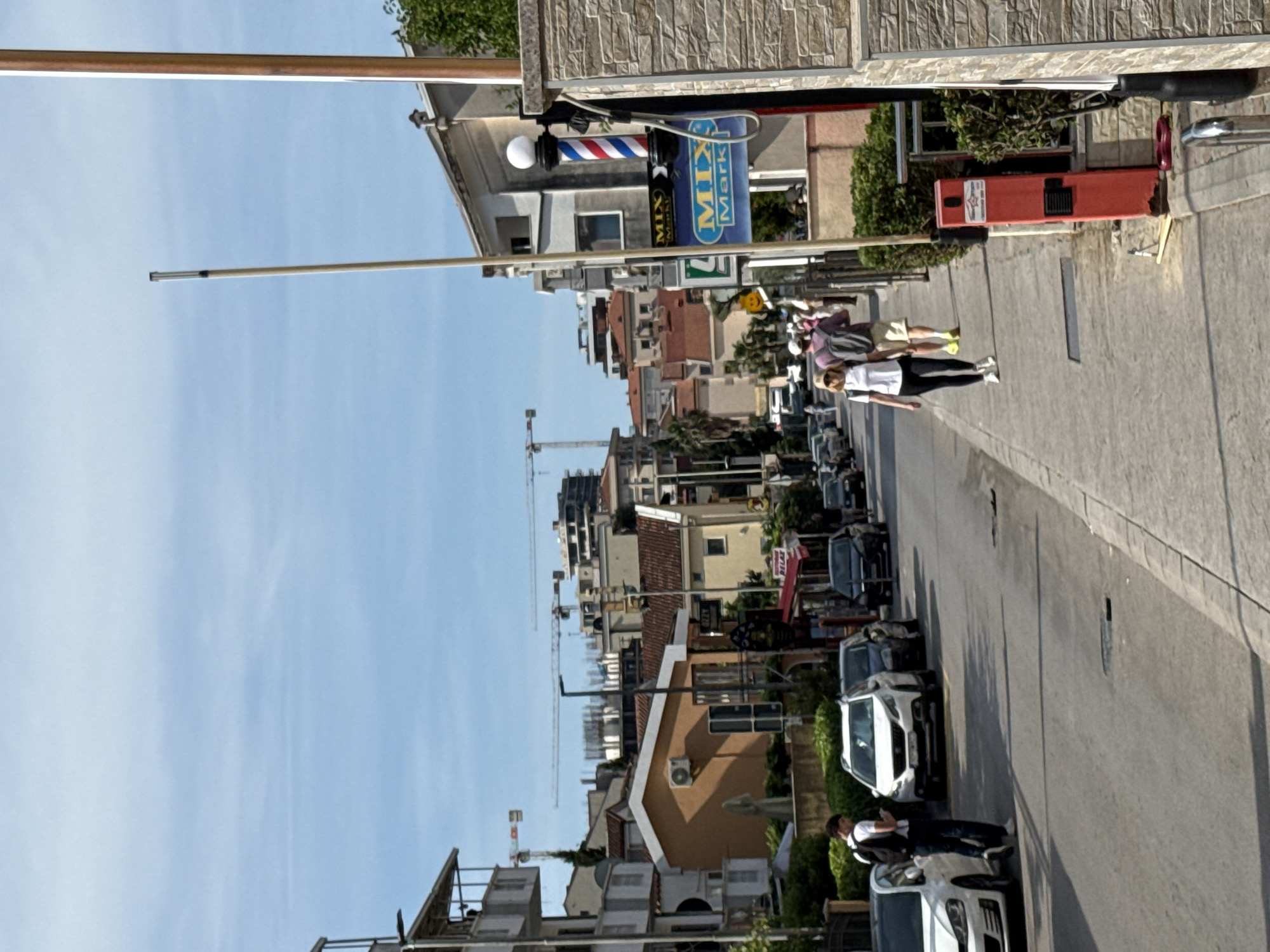 The image shows a street scene in Budva, Montenegro, captured on a sunny day. The street runs from the bottom to the top of the frame, lined with buildings on both sides. Parked cars are visible along the right side of the street. Two people are walking on the sidewalk on the right side of the street. A barber shop sign with a traditional rotating pole and a "Mix Markt" sign are visible near the top of the frame. In the distance, construction cranes dot the skyline amongst the buildings. The sky is blue with some wispy clouds. The scene suggests a typical urban environment.