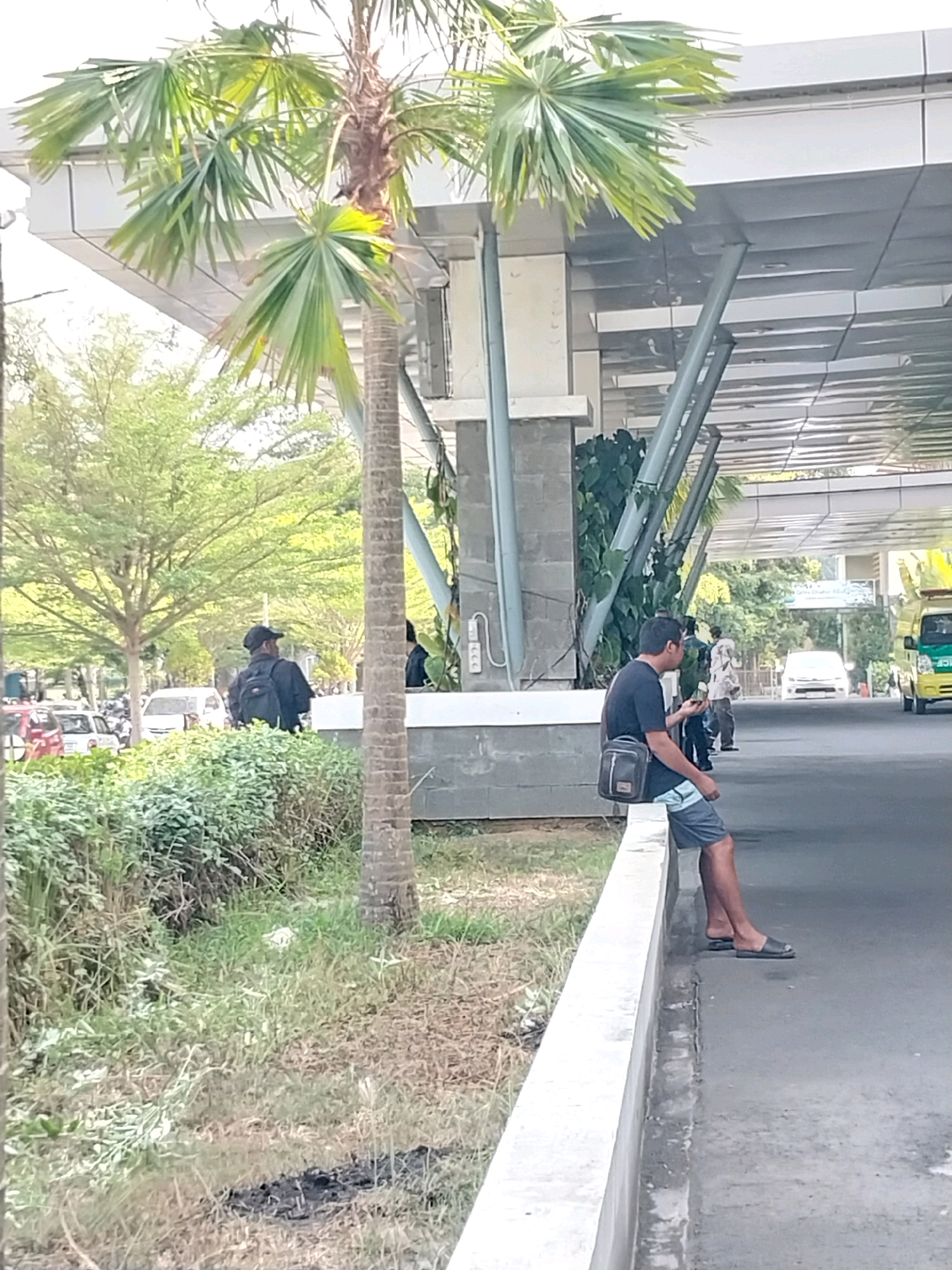 The image depicts an outdoor scene in Wates, Indonesia, likely near a public building or transportation hub, characterized by modern architecture. A man, wearing a black t-shirt, grey and blue patterned shorts, and black slip-on sandals, is seated on a low concrete barrier. He has a black shoulder bag with a strap across his body. The man is holding a mobile phone in his right hand and looking at it.
In the background, a palm tree stands prominently in the foreground. Behind the man, several vehicles, including cars and a bright yellow van, are visible on a paved road. Other individuals are present in the background, some appearing to be walking. The environment includes lush green foliage and trees. The overall setting suggests a well-maintained public space with some pedestrian and vehicular activity.