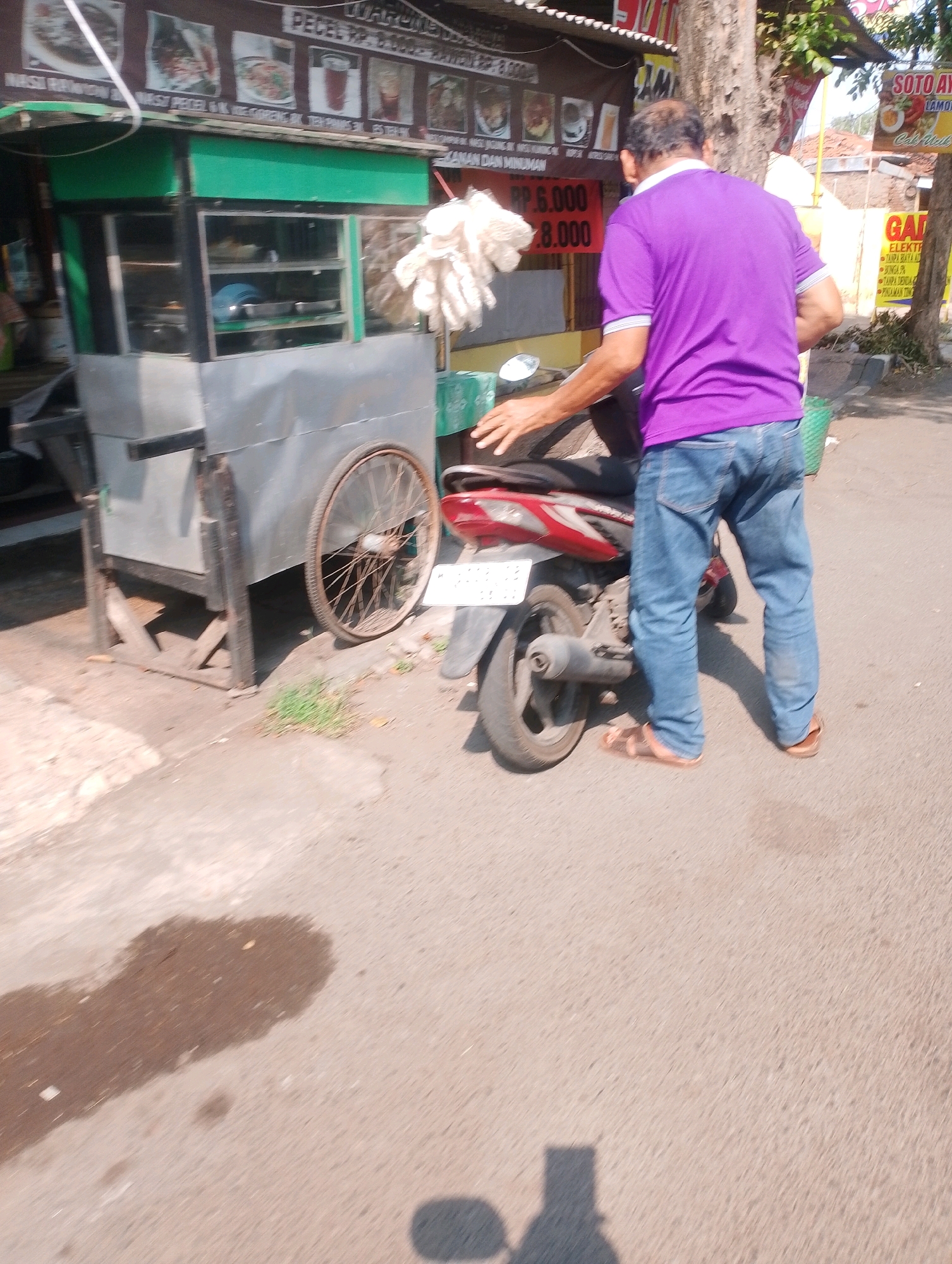 A man wearing a purple shirt and blue jeans is standing next to a red motorcycle. He is partially obscured by the motorcycle and a food cart. The food cart is made of metal and has a green awning. Behind the food cart, there is a sign with pictures of food. The scene appears to be in an outdoor market or street. There are trees and buildings in the background, suggesting an urban environment. The ground is paved with asphalt.