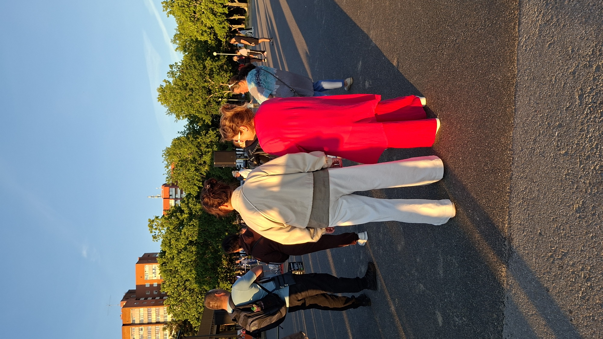 In the city of Tallinn, Estonia, a group of people are gathered in an outdoor area with a paved ground and trees in the background. Two women are in the foreground, facing away from the camera. The woman on the left wears a light-colored jacket and white pants, while the woman on the right is dressed in a bright red outfit. Several other individuals are visible in the mid-ground and background, some appearing to be talking or observing. A sound speaker is set up, suggesting a possible event or gathering. The scene is illuminated by the warm glow of sunset, casting long shadows. In the distance, multi-story buildings are visible above the trees.