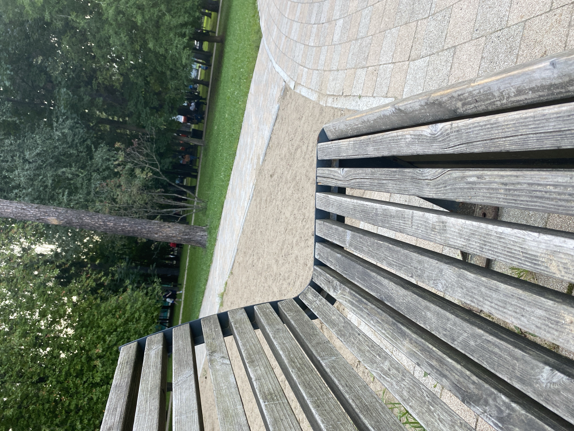 This is a photograph of a wooden park bench in Balashikha, Russia, taken at a low angle. The bench is weathered and grey, with visible grain and cracks in the wood. Behind the bench, a sandy area transitions into a paved pathway. Further back, a lawn area is visible with several trees and some people in the distance. The trees are lush and green, indicating it is likely summer or early autumn. The overall scene depicts a public park setting.