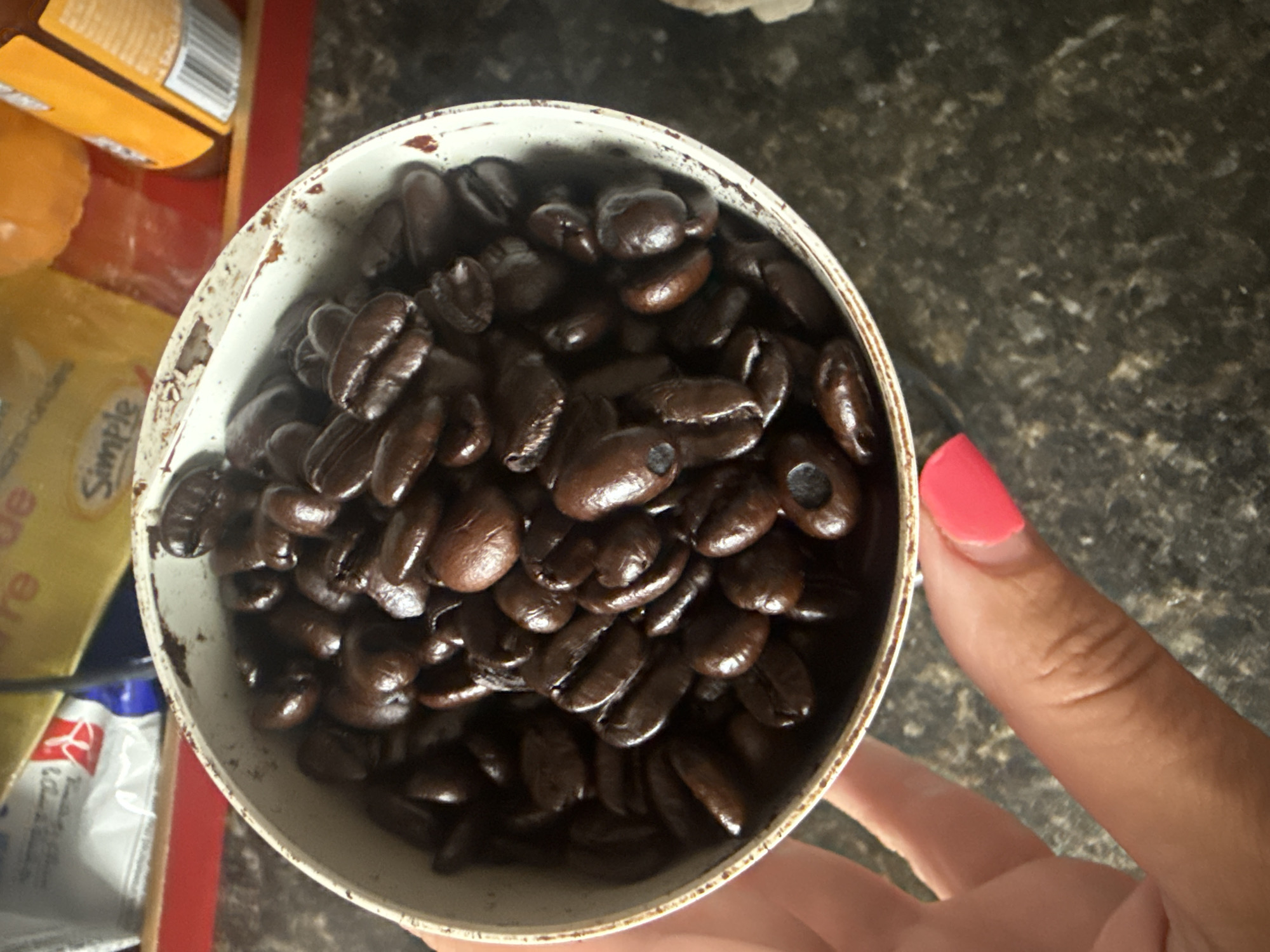 A close-up, high-angle shot shows a white bowl filled with dark roasted coffee beans. The bowl has a worn, chipped rim with a metallic edge. A person's hand, with bright pink nail polish on the index finger, is visible on the left side of the frame, holding the bowl from the side. The background is out of focus and appears to be a countertop with speckled patterns. On the counter, there are out-of-focus items including what looks like a yellow box with text and a dark orange bottle. The scene is indoors, possibly a kitchen or pantry. There is no information in the image to determine the location in Calgary, Canada.