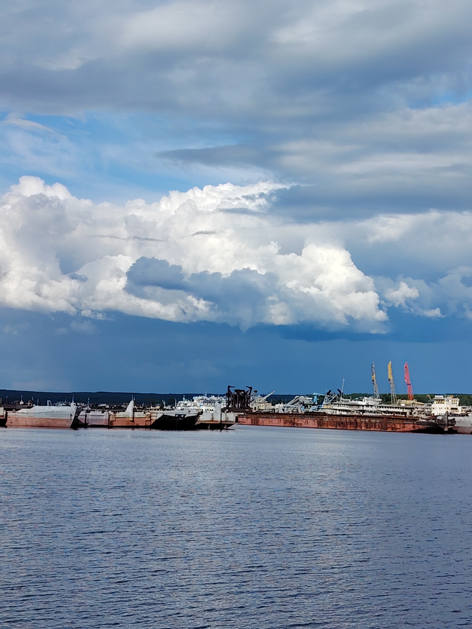 The image depicts a harbor scene in Chaikovsky, Russia, showing several ships and barges moored along the waterfront. The water is calm with slight ripples. In the background, a line of ships and barges are visible, some appearing old and possibly decommissioned. Two red and yellow cranes stand out among the vessels. The sky is dramatic with a mix of blue sky and heavy, dark clouds, suggesting possible inclement weather. The overall atmosphere is industrial and somewhat desolate.