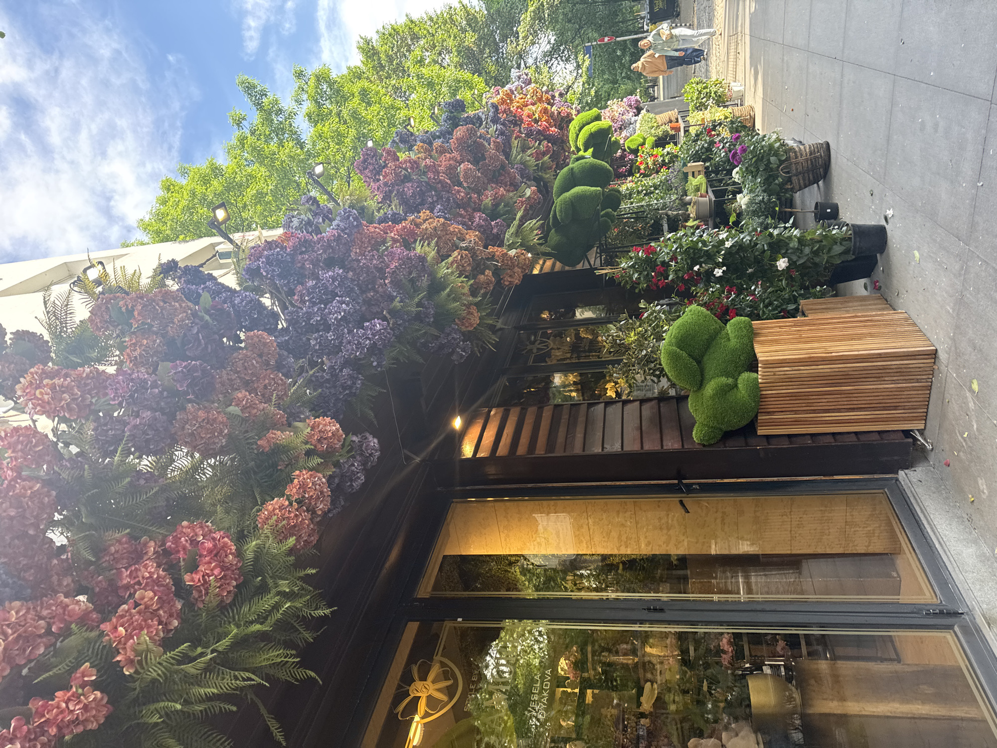 The image captures the storefront of a business adorned with an abundance of artificial flowers and greenery. The facade of the building is dark wood, and large glass windows provide a view into the interior. Above the windows, an elaborate display of artificial flowers in various colors (reds, purples, oranges) cascades down. Additionally, there are two topiary-like structures of green moss. A wooden bench is positioned outside the entrance.
The image is a street-level view, with a sidewalk visible on the right side of the frame. Potted plants are arranged along the sidewalk.
The sign on the window includes the words "VESELA BRAYAKOVA" The business appears to be in Sofia, Bulgaria. In the upper right corner, two pedestrians are walking away in the distance.