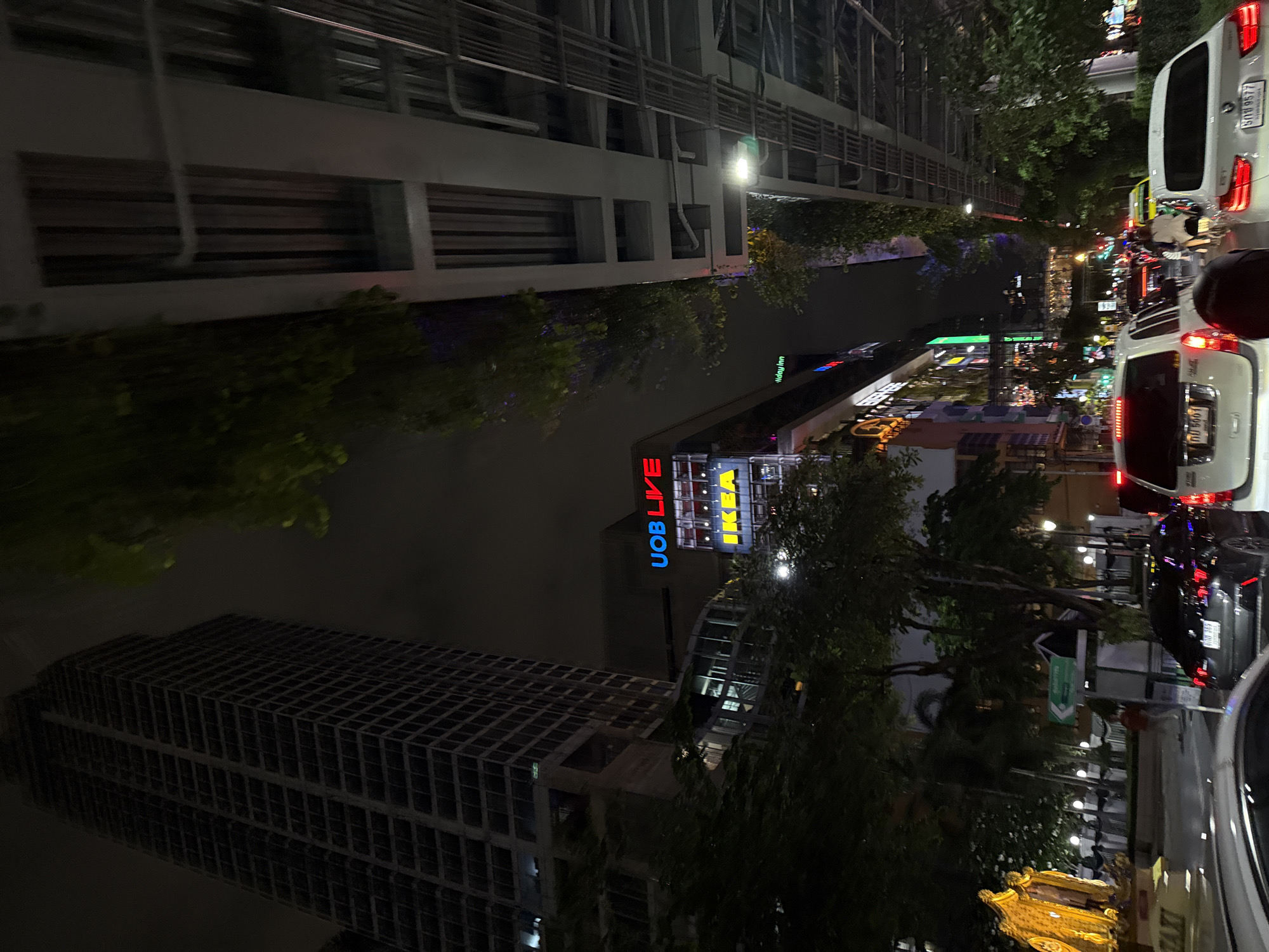 The image captures a nighttime scene on a city street, likely in Bangkok, Thailand, given the presence of recognizable landmarks and traffic signs. The perspective is low, suggesting the photo was taken from inside a vehicle.
Dominating the left side of the frame is a towering, dark skyscraper, its numerous windows creating a grid-like pattern against the night sky. To its right, a multi-story building displays prominent illuminated signage for "UOB LIVE" and "IKEA," indicating a commercial area. Further down the street, other illuminated signs and building facades suggest a bustling urban environment.
The street itself is congested with traffic. Several cars are visible, including white SUVs and a dark sedan. Red taillights of vehicles create streaks of light, emphasizing the slow-moving or stopped traffic. A yellow taxi is also present in the distance. Amidst the vehicles, a person on a motorcycle can be seen.
Trees and foliage are scattered throughout the scene, with some branches obscuring parts of the buildings and sky. An elevated structure, possibly an overpass or a large building's exterior, runs across the upper right portion of the image, supported by columns and featuring exposed conduits and pipes.
On the far left, a large, golden statue of figures is illuminated, standing next to a yellow taxi with the word "TAXI" visible. The overall atmosphere is one of a vibrant, active city at night, with a constant flow of traffic and illuminated structures.