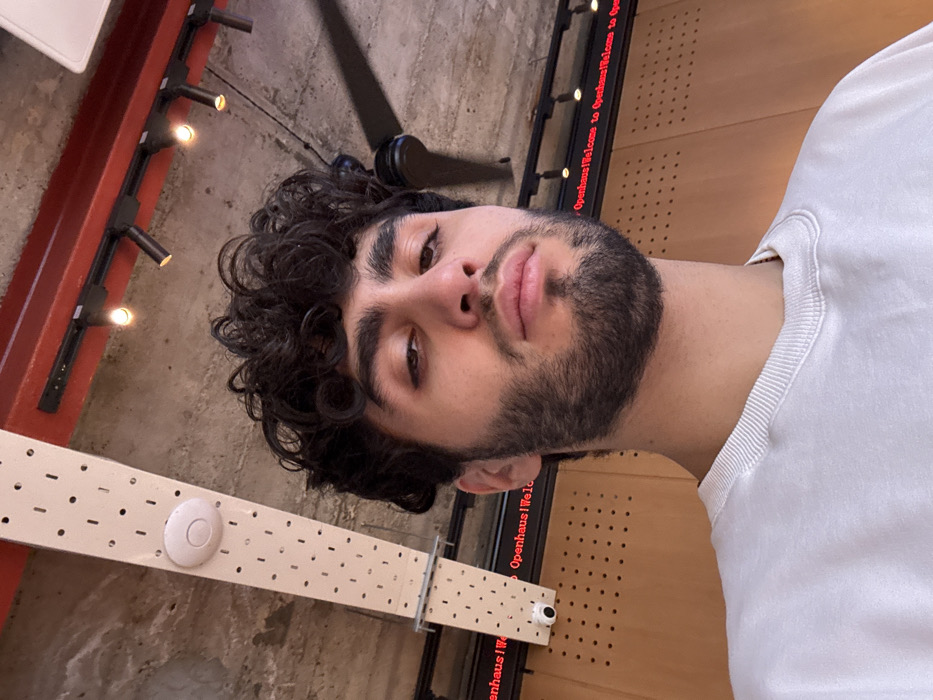 A young man with dark, curly hair and a beard stares directly at the camera. He wears a white t-shirt. Behind him, a ceiling features industrial elements like metal beams with lights and a perforated white structure. "Openhaus Welcome to Openhaus Welcome to" is displayed on a digital sign. The image is rotated, giving a tilted perspective.
