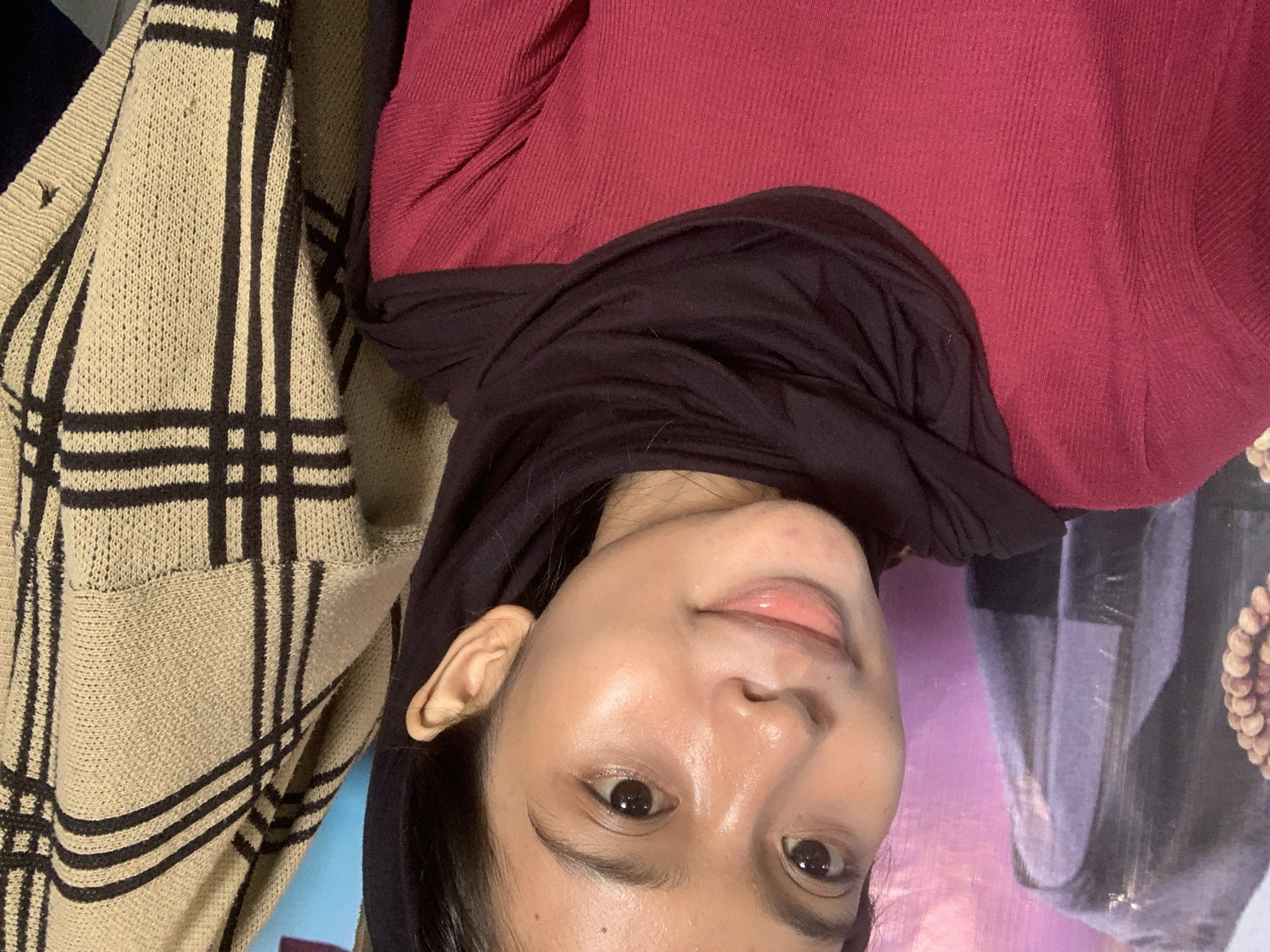 A young woman with light brown skin and dark brown eyes is looking directly at the camera in a close-up shot. She has on a dark purple hijab and a red long-sleeved shirt. Her face is slightly angled to the left, and her lips are parted. Her skin appears dewy and there are subtle highlights on her forehead and cheeks. A beige cardigan with a black plaid pattern is visible on the right side of the frame. In the background, on the left, a portion of a large poster with purple and black elements is visible. The image appears to have been taken indoors, likely in a residential setting within the city of Sungai Meriam, Indonesia.