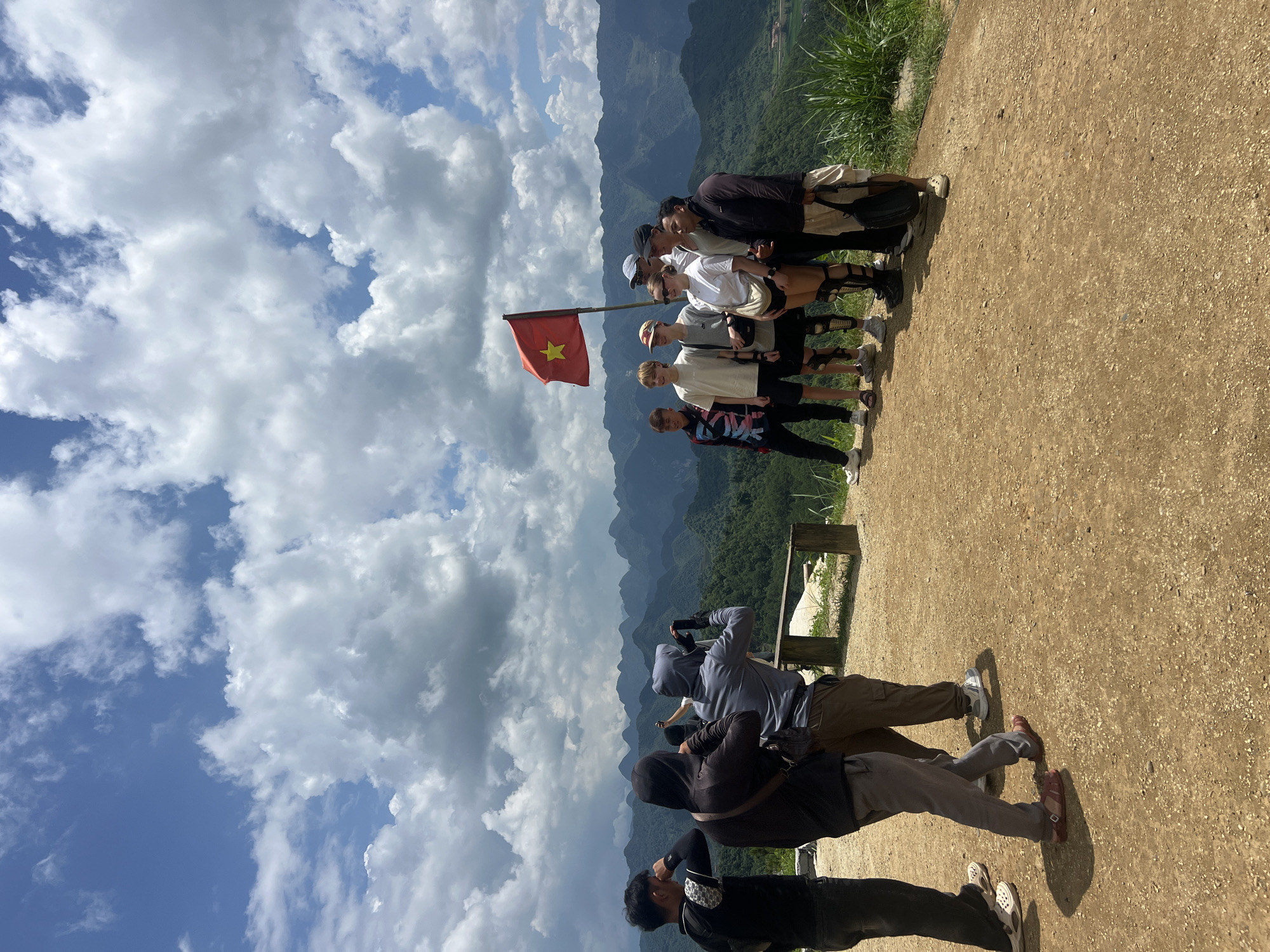 The image shows a group of eight people posing for a photograph on a dirt overlook with a mountainous landscape in the background under a cloudy sky. A Vietnamese flag is planted to the right of the group. Three people on the left of the frame are facing away from the camera, with two of them appearing to take photos. The remaining five people are facing the camera, posing for a picture. The individuals are a mix of genders and ages, wearing casual clothing. The environment is characterized by rolling green hills covered in lush vegetation, suggesting a rural or natural setting. The ground is covered in light brown dirt with scattered gravel. A wooden bench is visible to the left of the group, partially obscured by some of the individuals. The sky is filled with large, fluffy white clouds, with patches of blue sky visible. The scene suggests a tourist or sightseeing location, likely in Quản Bạ Commune, Vietnam, known for its karst topography.