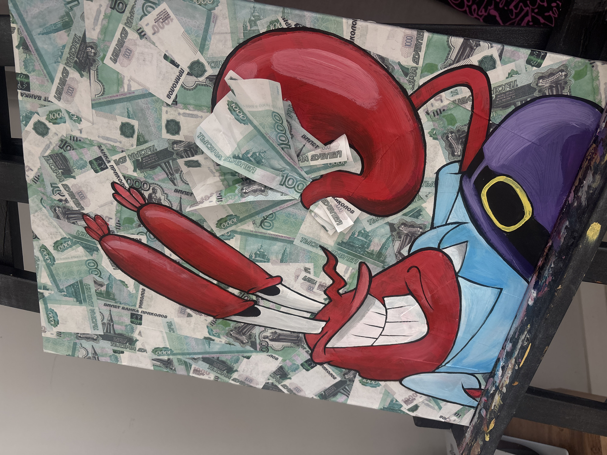 The image is a painting on canvas, displayed on a black easel. The painting features a character resembling Mr. Krabs from SpongeBob SquarePants, painted in a cartoonish style. The character is red with blue shirt and purple pants. The background of the painting is made of what appears to be Russian 1000 ruble banknotes. The easel is positioned against a plain, light-colored wall. Some paint splatters are visible on the horizontal bar of the easel. The overall setting suggests an indoor art display or studio. There is no indication of the specific location within the city of Krasnoyarsk, Russia.