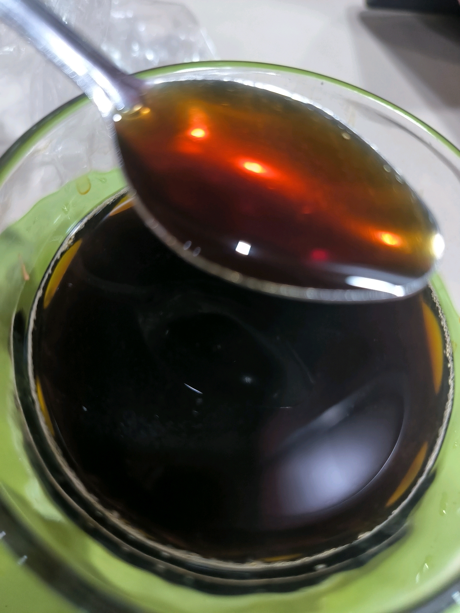 A close-up shot shows a spoonful of dark brown liquid being held over a glass containing a similar dark liquid, possibly coffee or tea. The spoon is metallic, and its surface reflects light. The glass is green and has a smooth interior. The lighting in the image is bright, creating reflections and highlights on the liquids and the spoon. The background is blurred and appears to be a light-colored surface, possibly a table. There are no people visible in the image. The scene is indoors and does not provide any specific visual cues to determine its location within Banda Aceh, Indonesia.