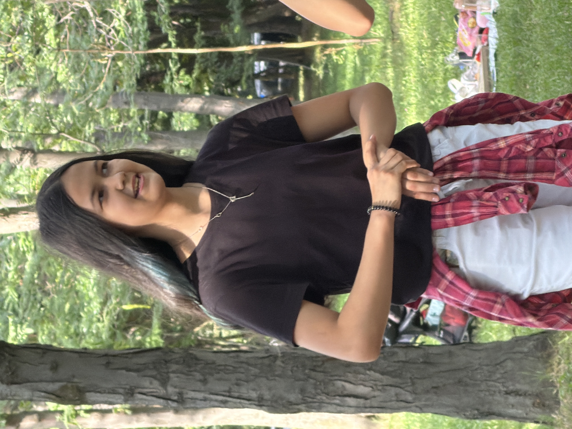 A young girl with long, dark hair and streaks of blue is standing in a park. She is wearing a black t-shirt, white shorts, and a red and white plaid shirt tied around her waist. Her hands are clasped in front of her. In the background, there are trees and a picnic spread on the grass.