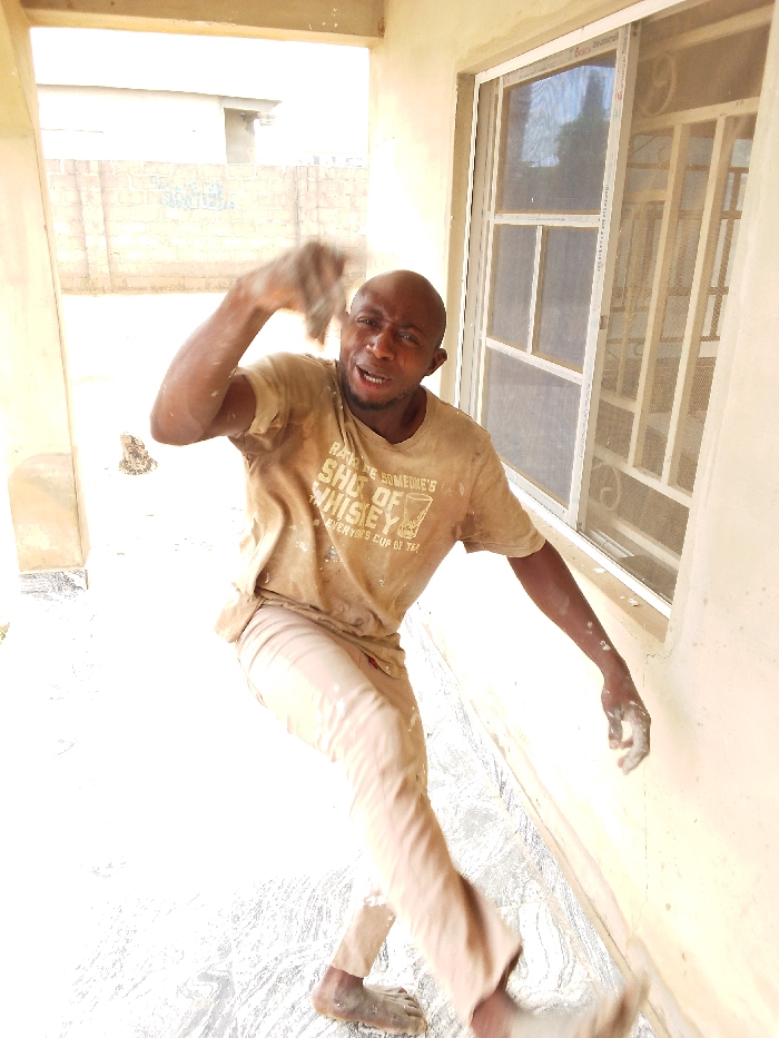 In Ajaokuta, Nigeria, an adult male, bald and with facial hair, is captured mid-action on what appears to be a building's porch or veranda. He is barefoot, wearing a light brown, paint-splattered t-shirt that reads "RATHER BE SOMEONE'S SHOT OF WHISKEY THAN EVERYONE'S CUP OF TEA," and light brown trousers. White paint or plaster dust covers his clothing, arms, hands, and feet. His left arm is raised, hand clenched, holding a white-stained rag, while his right arm is extended downwards. His face shows an open-mouthed expression of exertion or surprise. To his right, a white-framed window with internal security grilles and mesh is set into a light-colored wall, which also shows signs of renovation. The light-colored floor beneath him is similarly splattered with white material. In the background, through an opening, an outdoor area features a light-colored block wall with faint graffiti, a distant building, and a pile of dark material on the ground, all under a bright sky. The scene suggests active construction or renovation work.