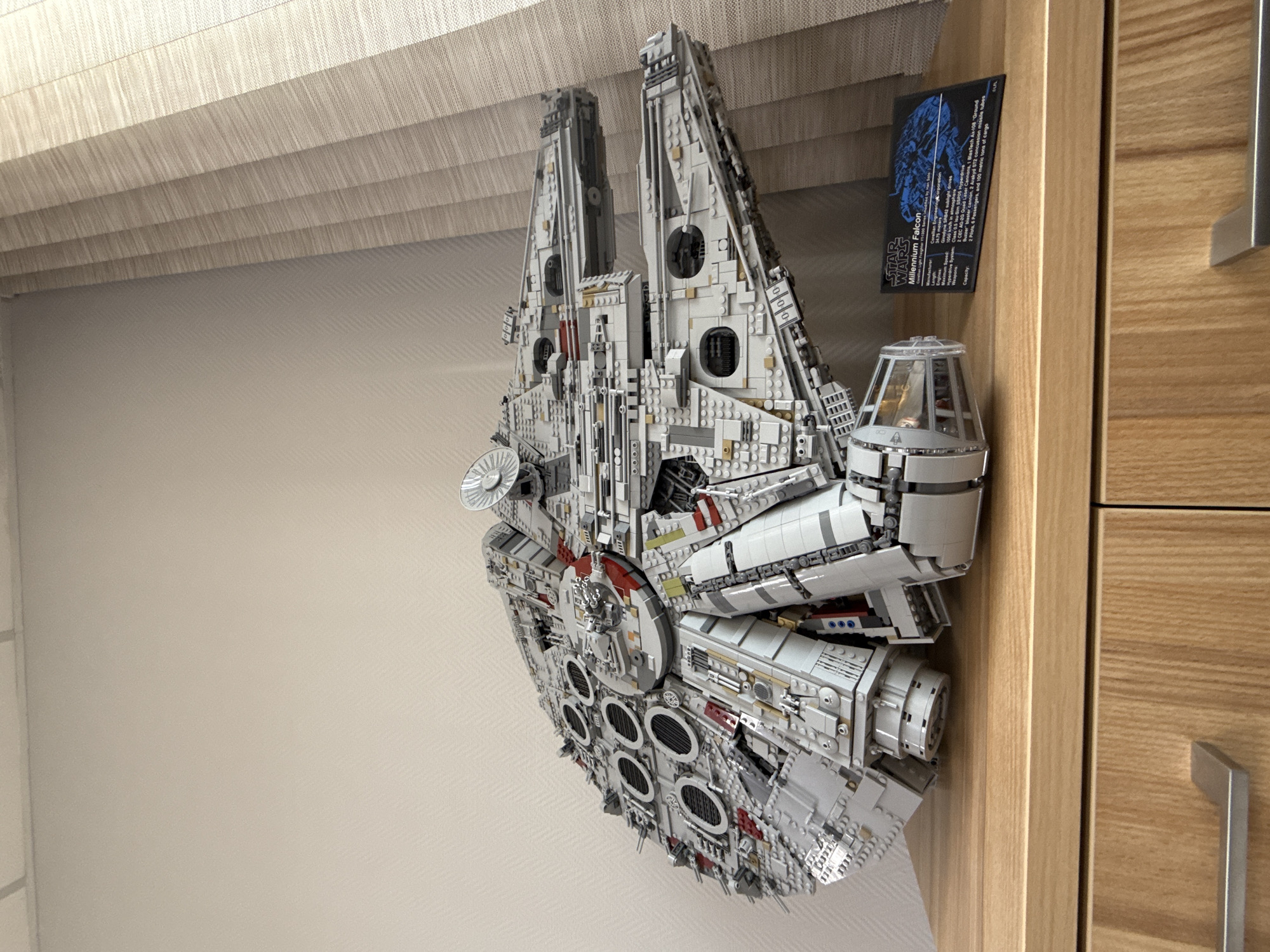 An indoor scene features a large, intricately detailed LEGO model of the Star Wars Millennium Falcon, prominently displayed. The model, primarily constructed from light and dark grey bricks with subtle red and yellow accents, appears to be a collector's edition given its scale and complexity. It is positioned against a light-colored, textured wall and oriented as if angled slightly upwards.
In the upper left portion of the image, a light-colored pleated blind or curtain is visible, suggesting a window or overhead light source. To the right, a section of a wooden cabinet or furniture piece is present, featuring a silver metal handle.
Adjacent to the cockpit section of the Millennium Falcon model, a black rectangular plaque with white text and a blue graphic is displayed. The text on the plaque identifies the subject as the "STAR WARS Millennium Falcon" and lists various specifications, including: "Manufacturer: Corellian Engineering Corporation," "Sublight drives: Girodyne SRB42 sublight drives," "Speed: 1000 km/h in atmosphere," "Hyperdrive System: Class 0.5," "Weapons: 2 CEC AG-2G quad laser cannons, 1 BlasTech Ax-108 'Ground Buzzer' concussion missile launcher," and "Capacity: 2 Pilots, 6 Passengers, and 100 metric tons of cargo." The scene suggests a personal display of a cherished collectible. No specific time of day or weather conditions are discernible, and there are no people visible. The image provides no visual cues for the location of Krasnoyarsk, Russia.