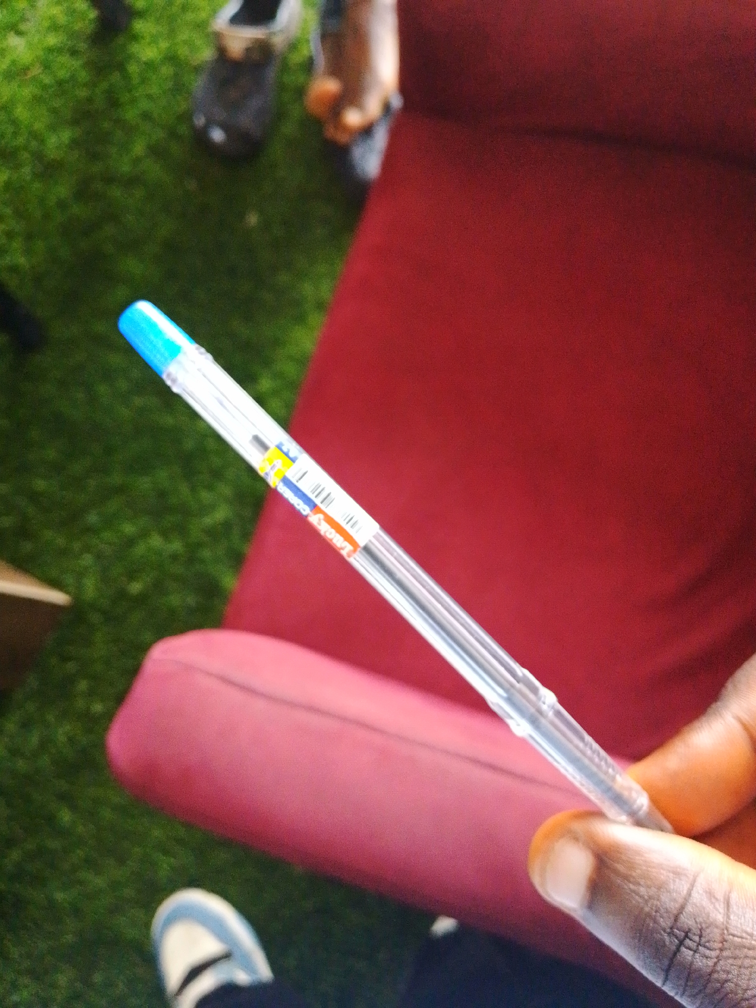 A clear plastic pen with a blue cap is held in a person's right hand, which is in the foreground. The pen has a label that reads "Lucky Corra" in orange letters. The background shows a red chair or sofa with a textured surface, and a grassy area. In the far background, blurred figures and footwear are visible. This scene is depicted in Barikin Jirga Summa, Nigeria.