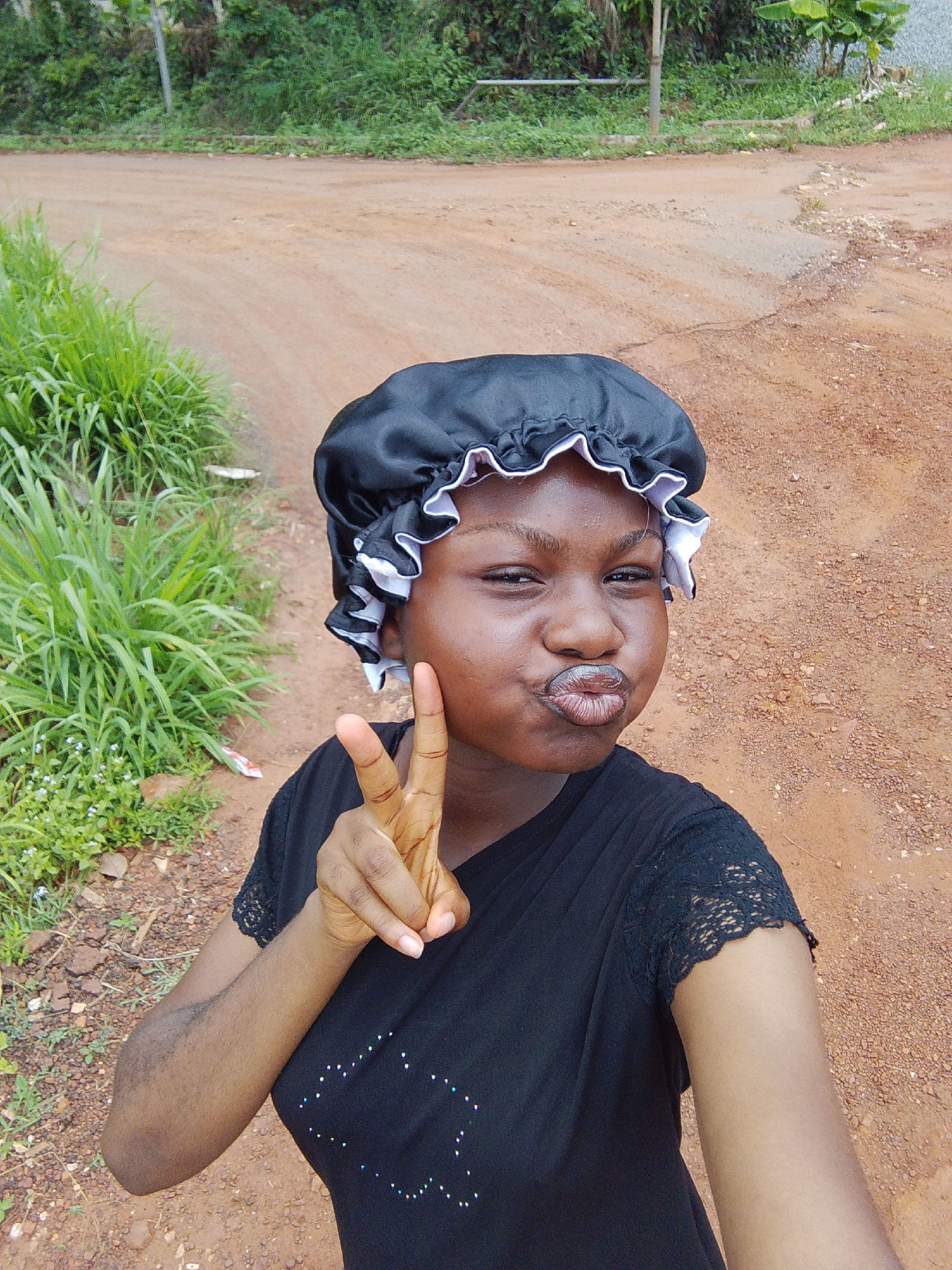 A young woman is posing for a selfie outdoors on a dirt road. She is wearing a black satin bonnet with white ruffles and a black t-shirt with lace sleeves. She is making a peace sign with her right hand and puckering her lips. There is vegetation to the left of her and a dirt road in the background. Based on the given location, the photo was likely taken in Nsukka, Nigeria.