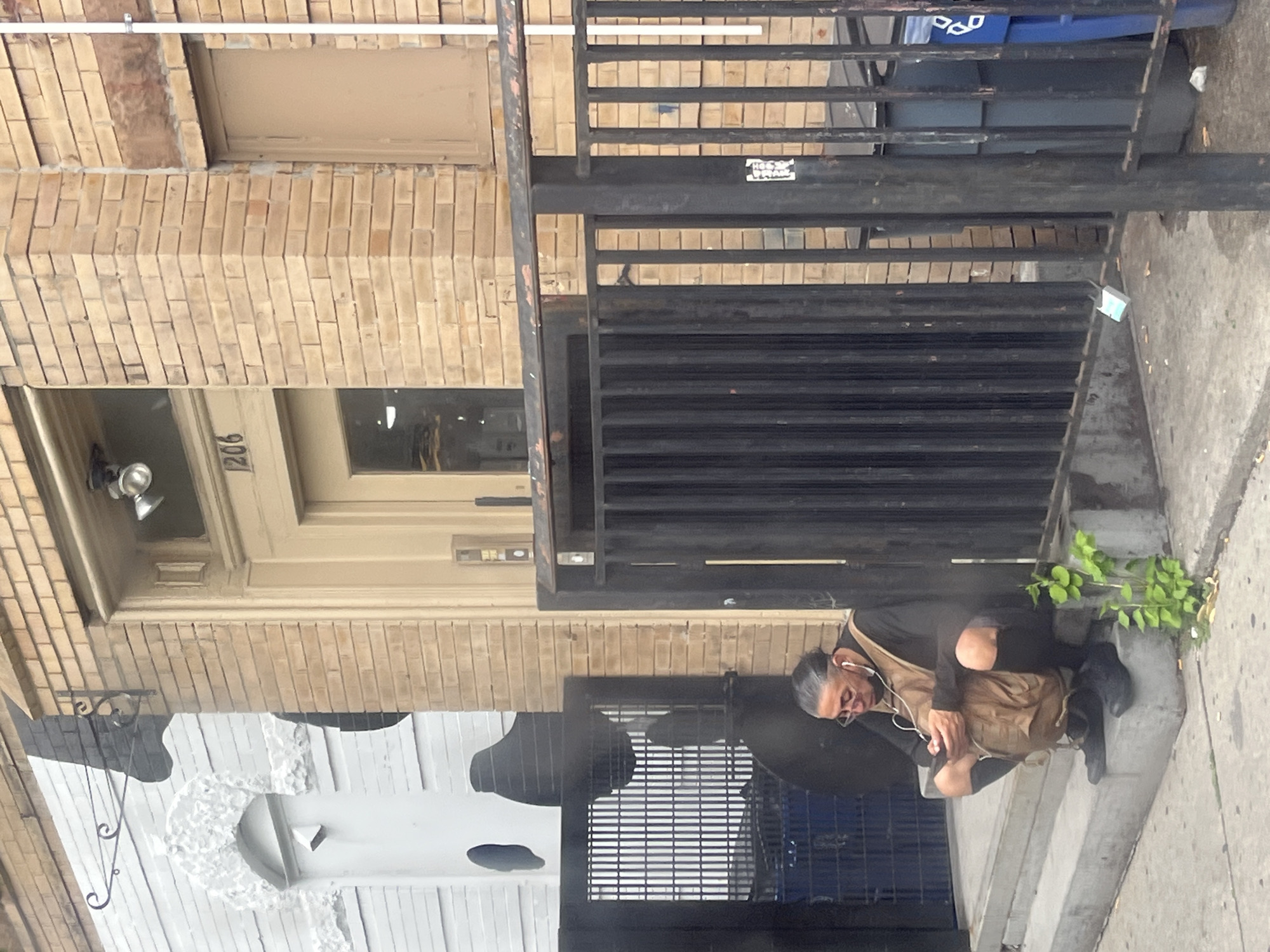 The image depicts a street scene in New York City, likely during the daytime, given the lighting. A person is squatting on a set of concrete steps next to a building. The building's facade is made of light-colored brick, with a door labeled "1206" visible. There's a black metal gate in front of a section of the building.
The person is wearing dark clothing, including ripped pants, a black long-sleeved shirt, and black boots. They have a tan messenger bag slung over their shoulder and are wearing earbuds. Their facial expression is neutral or perhaps weary. Small green plants are growing from a crack in the concrete near the steps.
In the background, through the metal gate, a blue recycling bin and a dark gray trash can are visible. The ground is paved with concrete. The overall atmosphere appears to be urban and somewhat gritty, a common characteristic of street scenes in many parts of New York. No specific event is clearly taking place, other than the individual resting on the steps.