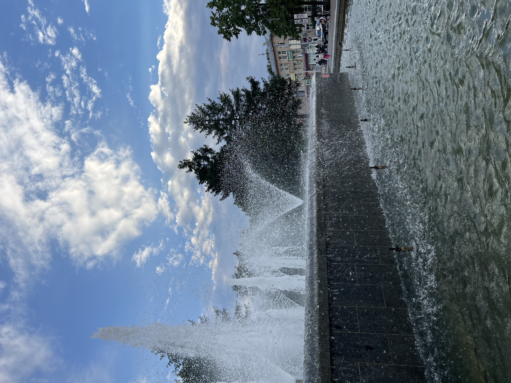 This media file captures a view of a fountain in Chaikovsky, Russia. The scene is dominated by a large, dark-tiled fountain with water spraying upwards and outwards, creating a dynamic and refreshing visual. Beyond the fountain, lush green trees frame a backdrop of buildings, suggesting an urban or park setting. The sky above is a vibrant blue, dotted with fluffy white clouds, indicating fair weather. Sunlight glints off the water droplets, highlighting the movement and energy of the fountain.
In the background, a street can be seen with a few people walking and some parked vehicles. The buildings appear to be multi-story structures, typical of a town or city. Some signs are visible on the buildings, though the text is not legible.
The activity taking place is the operation of a public fountain, likely a point of interest or recreation for visitors and residents. The overall mood conveyed is one of pleasant outdoor activity, with the cooling spray of the fountain offering a sense of respite on what appears to be a warm day. The composition, with the fountain in the foreground and the town in the background against a bright sky, creates a sense of depth and place. The time of day appears to be daytime, possibly late morning or afternoon, given the quality of light.