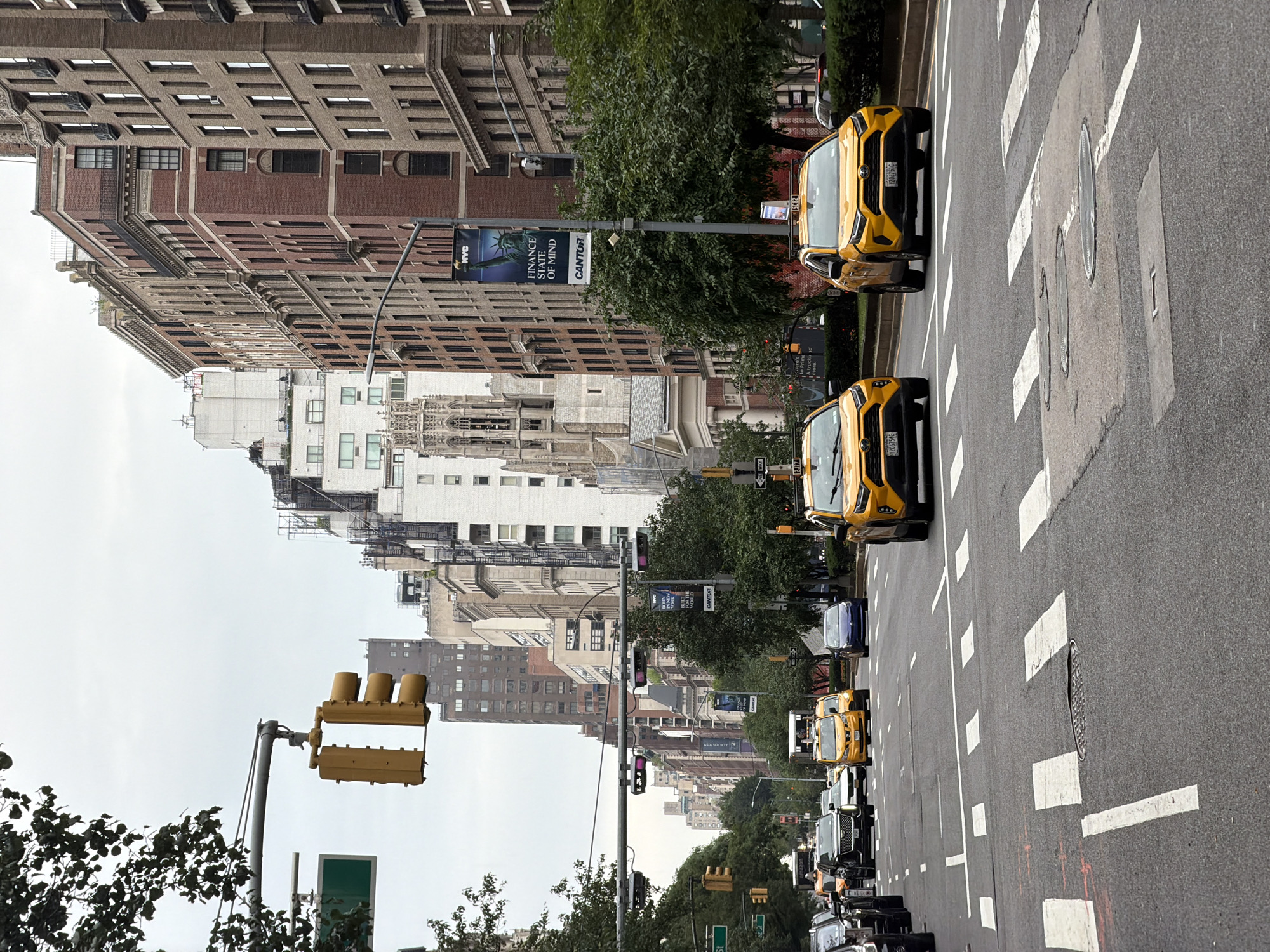 This is a medium-angle, eye-level, landscape photograph taken from the perspective of someone standing on a sidewalk or crosswalk, looking down a city street. The image is oriented vertically, with the top of the buildings appearing on the left side of the frame. The scene depicts a bustling street in New York City, with several yellow taxis driving down the road. The buildings on either side of the street are tall and made of brick and stone, characteristic of early 20th-century architecture. Trees line the sidewalks, adding greenery to the urban environment. A traffic light is visible in the foreground, and a pedestrian crossing is marked on the asphalt. The sky is overcast, suggesting a cloudy or hazy day. Banners are hung on poles along the street, with one clearly displaying the text "NYC FINANCE STATE OF MIND CANTOR". Other visible text includes "ONE WAY" and license plate numbers like "2J77", "5C82", "Y2067540", and "Y2067510". The overall impression is of a typical day in a major metropolitan area, with urban life in full swing.
