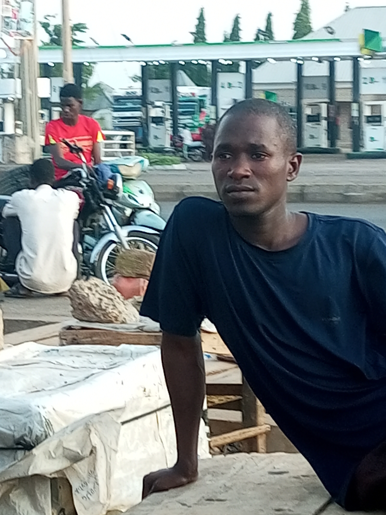 The image shows a man in a dark blue t-shirt sitting at an outdoor location in an urban setting. Behind him, there are two men, one in a red shirt and another in a white shirt, sitting on a motorcycle loaded with goods. In the background is a gas station. The specific location is Unknown, Unknown.