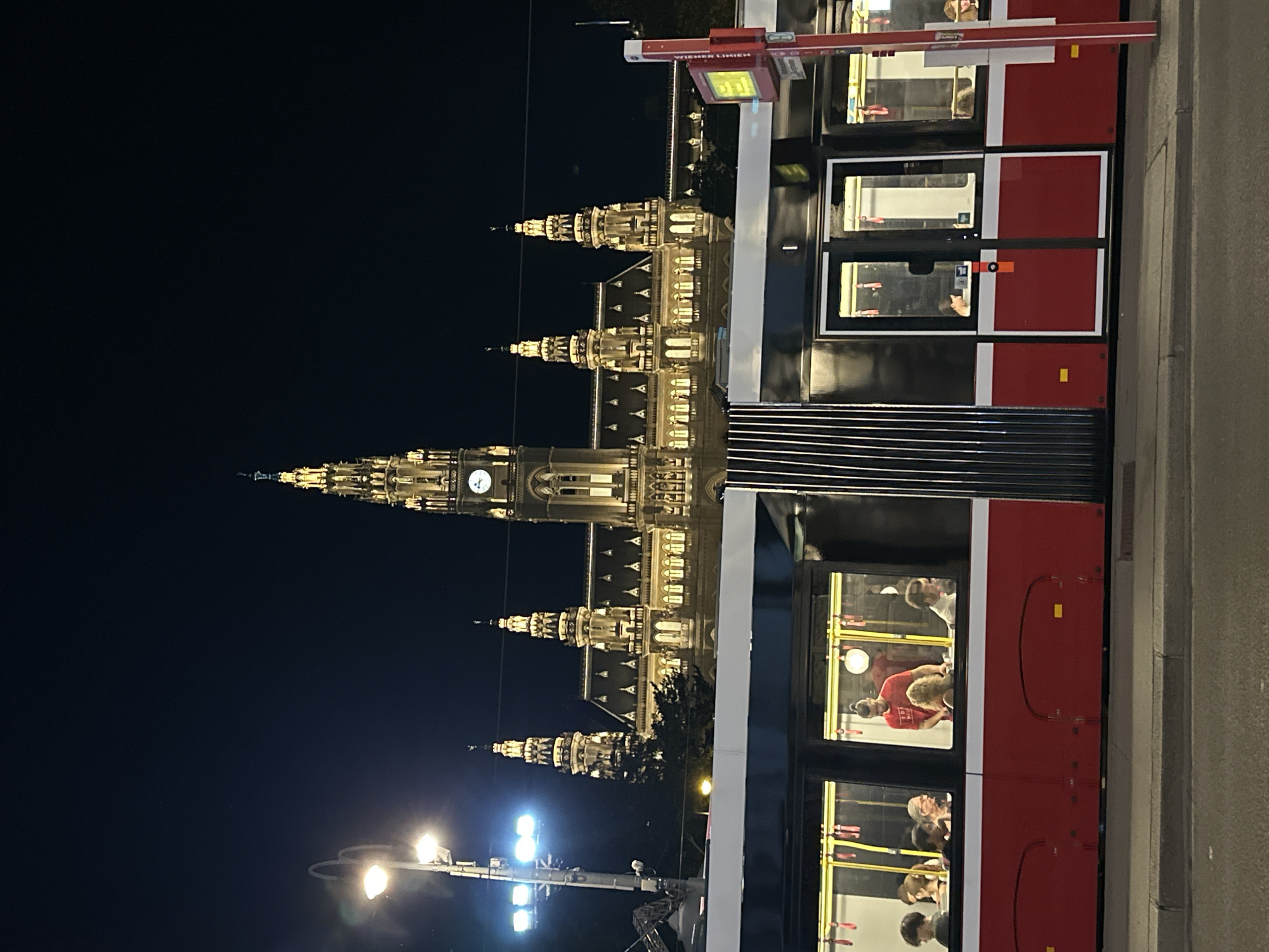 The image captures a night scene in Vienna, Austria, focusing on the Vienna City Hall (Rathaus) and a red and black tram passing in front of it. The City Hall is a large, ornate Gothic Revival building, brightly illuminated and detailed with spires, arched windows, and a prominent clock tower. The tram, with its distinctive red lower half and black upper section, has its windows revealing several passengers inside. The tram is stopped or moving slowly, positioned on a paved street. Streetlights are visible, casting a warm glow. The sky is dark, indicating nighttime. The overall impression is of a well-lit urban environment at night, with the historic architecture of the City Hall serving as a dramatic backdrop to the modern public transportation.