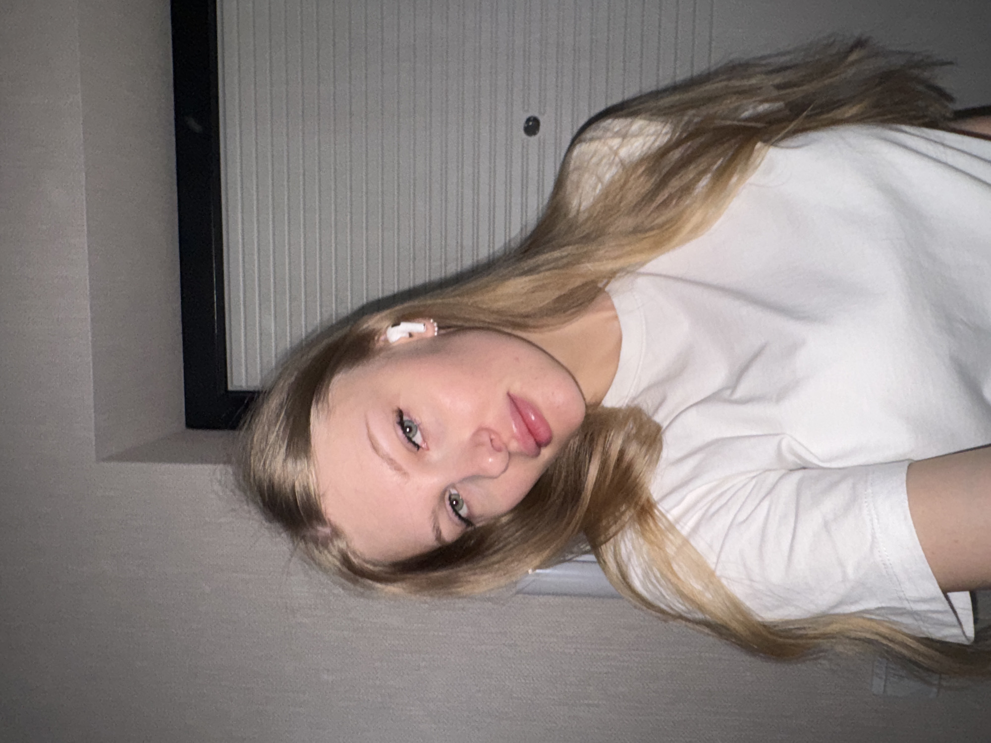 A young woman with long, light blonde hair and fair skin is posing for a selfie. She is wearing a white T-shirt and has white earbuds in her ear. She is inside a room, with a wall with vertical ribbed texture and a black-framed window or picture on the wall behind her. The specific location in Perm, Russia, cannot be determined from the image alone.