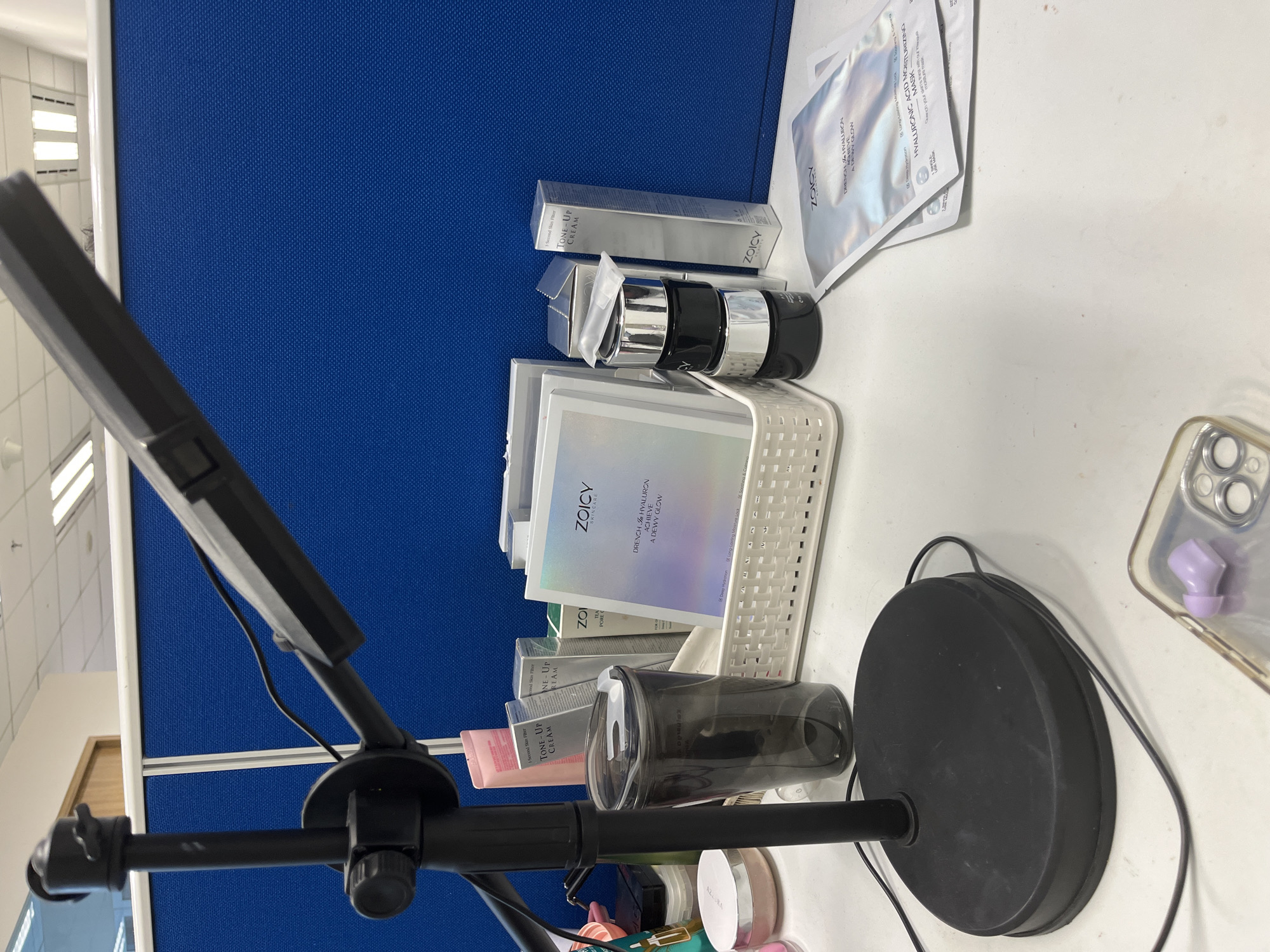 The image depicts a desk with various skincare products and a lamp in an office setting, possibly in South Jakarta, Indonesia. The desk is white and cluttered with items. In the foreground, a black desk lamp with an adjustable arm and head is prominent. To the right of the lamp, a dark gray tumbler with a lid is visible. Behind the tumbler, a white woven basket contains several skincare products, including boxes of "Tone-Up Cream" and a product from the brand "ZOICY" with the description "DRENCH 4% HYALURON ACHIEVE A DEWY GLOW." To the right of the basket are three black jars with silver lids stacked on top of each other. Further to the right, two silver foil packets of "ZOICY HYALURONIC ACID MOISTURIZING MASK" are laid out on the desk. In the background, a blue partition wall separates this workspace from another. The environment suggests a personal workspace within an office or a home office.