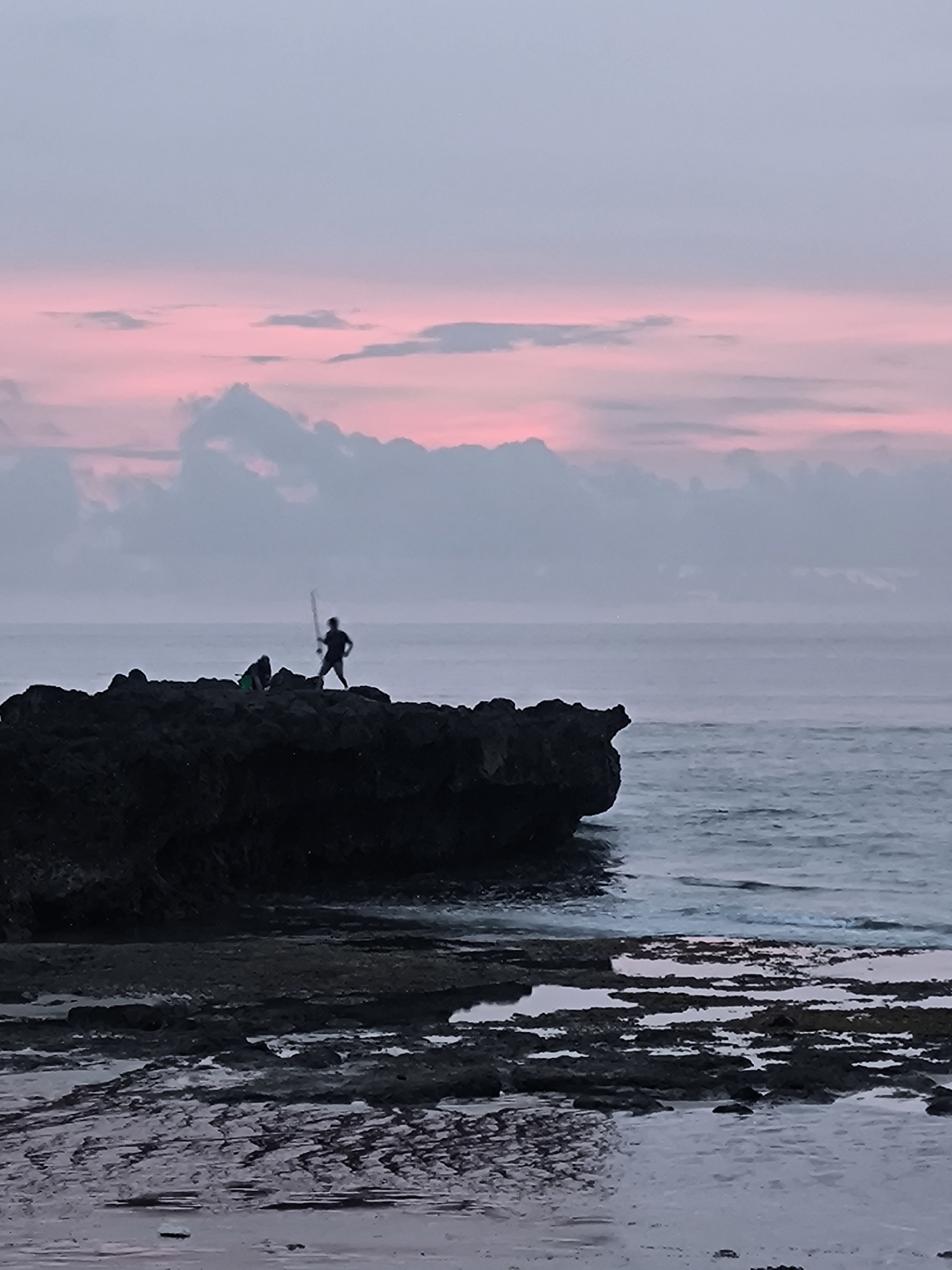 The image depicts a coastal scene in Canggu, Indonesia, at dusk or dawn, evidenced by the pink and grey sky. A dark, rocky outcrop is positioned in the middle ground, extending into the sea. Two figures are silhouetted against the sky, standing on the rocks. One is holding a long object, possibly a fishing rod or spear. The foreground features a wet, rocky beach area with shallow pools reflecting the sky. The sea is visible in the background, with gentle waves breaking.