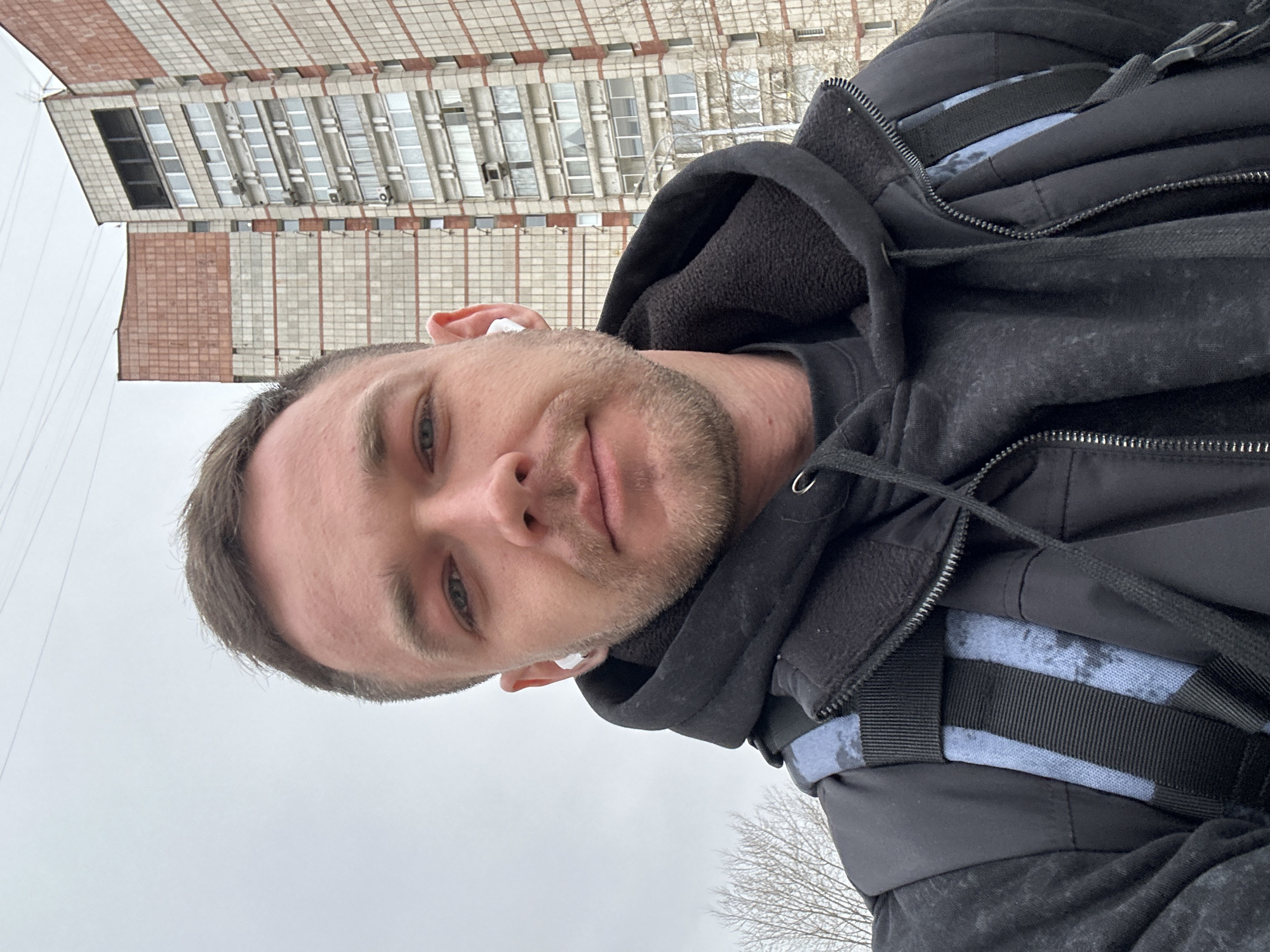 A selfie taken by a light-skinned male in Perm, Russia. He has short brown hair, blue eyes, and light stubble. He is wearing a black hooded sweatshirt and a black jacket with straps. He has white wireless earbuds in both ears. Behind him is a tall, older building with a brick facade and many windows. The sky is overcast. Bare tree branches are visible in the bottom right corner of the frame.