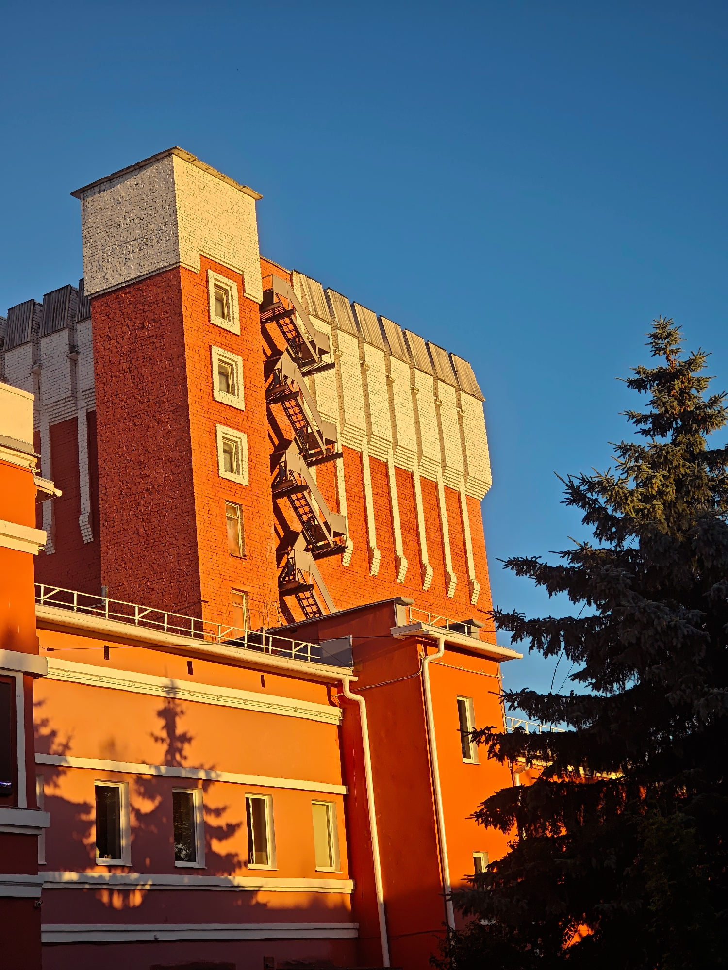This is a photograph taken from a low angle, looking up at the side of a building and a tree against a clear blue sky. The building is predominantly orange and red brick with white accents. There is a fire escape visible on the side of the brick section. The lower part of the building is painted a vibrant orange and features several windows. The shadow of a conifer tree is cast upon this orange section, creating an intricate pattern. The top section of the building is a tower-like structure, clad in white brick with red brick detailing at the base. A large conifer tree with dark green needles is positioned to the right of the building, partially obscuring it. The sunlight is hitting the building from the right, casting long shadows and highlighting the textures of the brickwork and the tree. The image appears to be taken during the golden hour, likely late afternoon or early morning, due to the warm lighting. The location is identified as Kimry, Russia, although no specific landmarks are visible to confirm this definitively within the frame. There are no people or animals visible in the image.