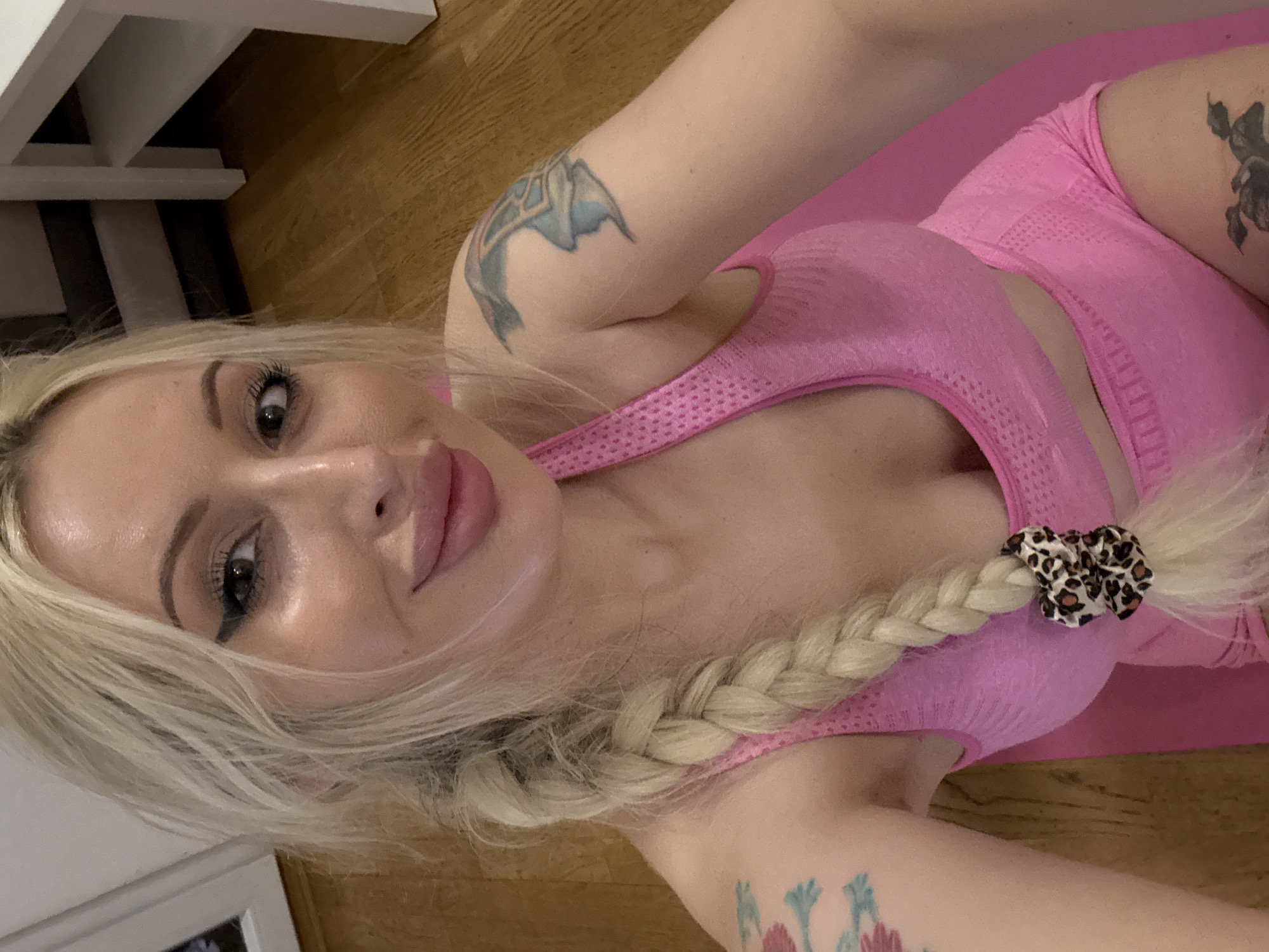 A blonde woman with a braid is posing for a selfie. She is wearing a pink sports bra and matching shorts. Her makeup is visible, with winged eyeliner and bold lipstick. She has tattoos on her left arm and leg, and a leopard print scrunchie in her hair. The background shows a wooden floor and a white table. The lighting suggests an indoor setting, possibly a home.
