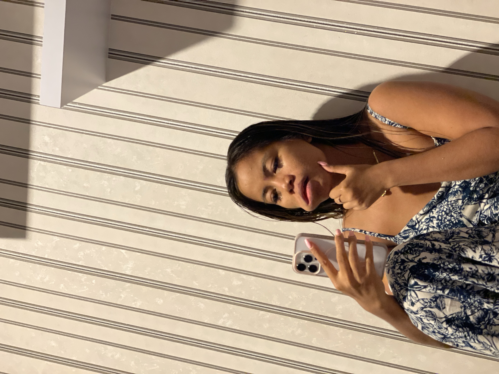 A woman is holding a smartphone, oriented horizontally, in front of her while posing for a photograph. She has dark hair, brown skin, and is wearing a blue and white floral patterned top. The phone is light-colored and has multiple camera lenses visible. She is touching her chin with one finger. The background is a wall with horizontal stripes. There is a small, white rectangular object visible in the top-left corner. The location is potentially in a building in Central Jakarta, Indonesia, though this is based solely on the user's query and not verifiable within the image itself.