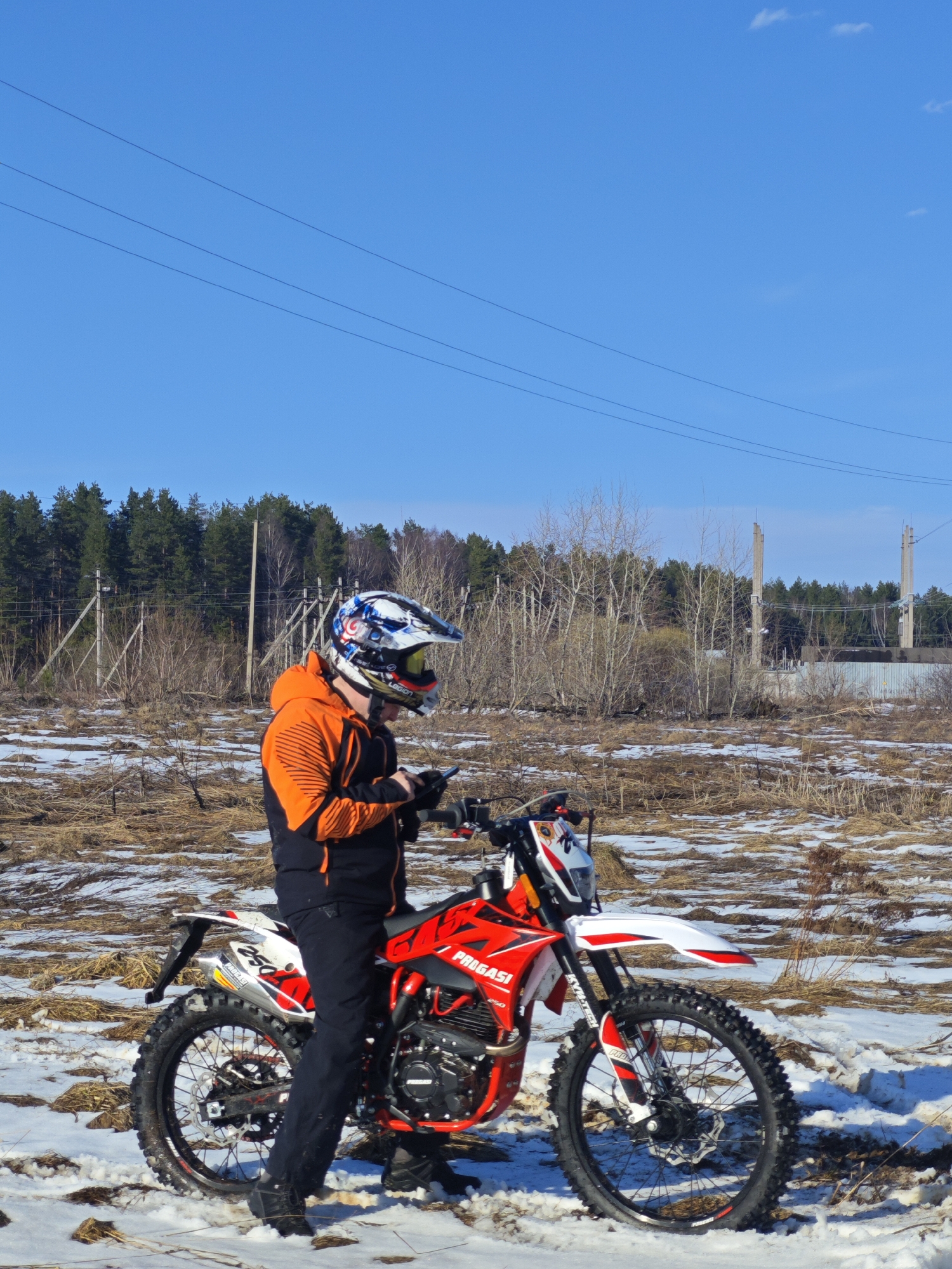A person wearing an orange and black jacket, black pants, and a multi-colored helmet is sitting on a red and white Progasi 250 dirt bike in a field of brown grass and patches of snow. The person is looking down at a handheld device. In the background, there is a line of evergreen trees, bare trees, a wire fence, and concrete structures resembling utility poles. The sky is a clear blue with a few faint clouds. Thin power lines stretch across the upper portion of the sky. Based on location information, the scene is in Прикамский, Russia.