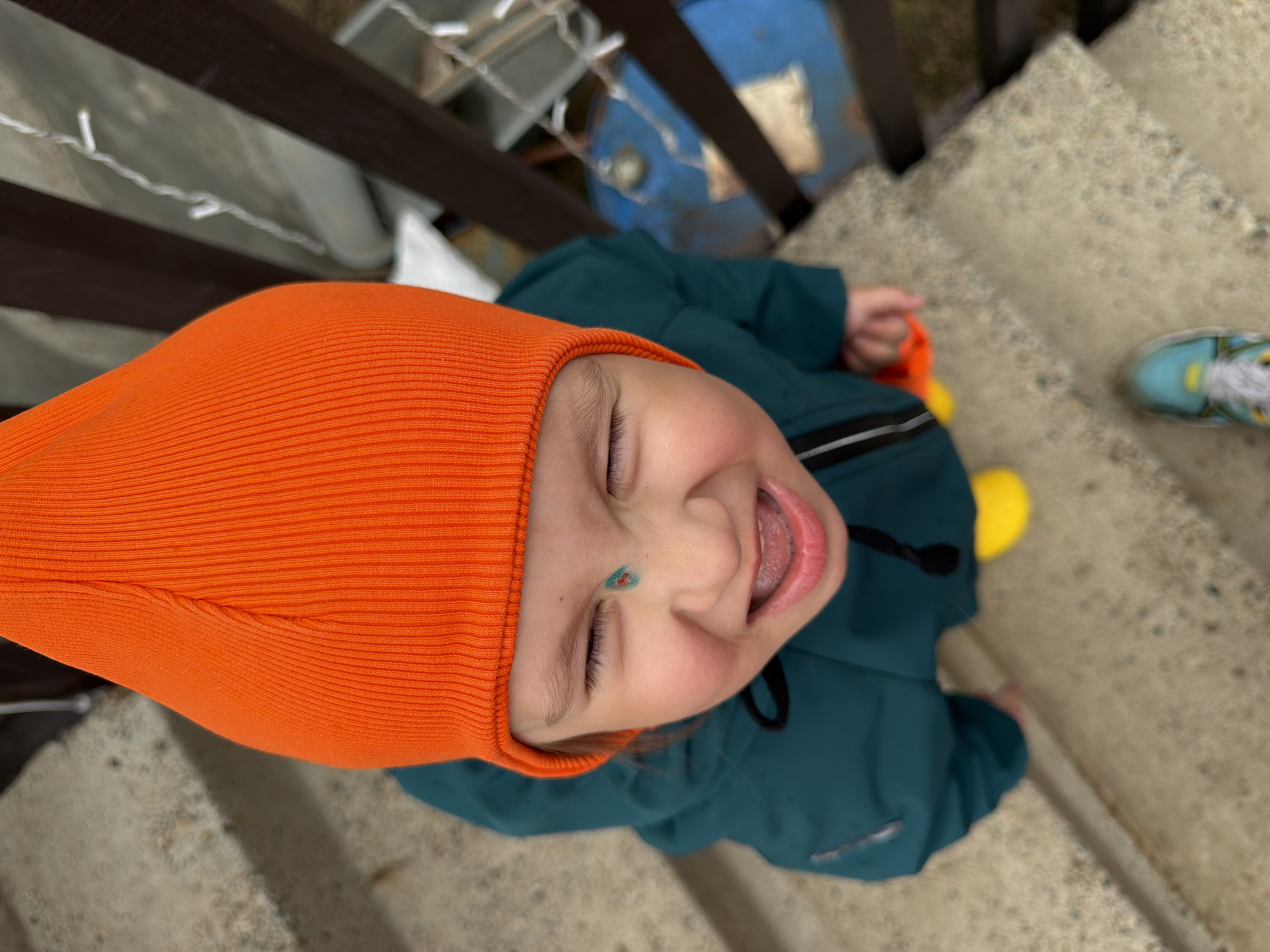 A child, facing upward, is wearing an orange hat and a teal jacket while sitting on concrete steps. The child has a green and blue mark on their nose, their eyes are closed, and their mouth is open. To the right of the child, a blue and yellow shoe is visible. A blue container is visible in the background through wooden railing. The location is likely in an urban area, potentially in Chaikovsky, Russia, but this cannot be confirmed with the available visual information.