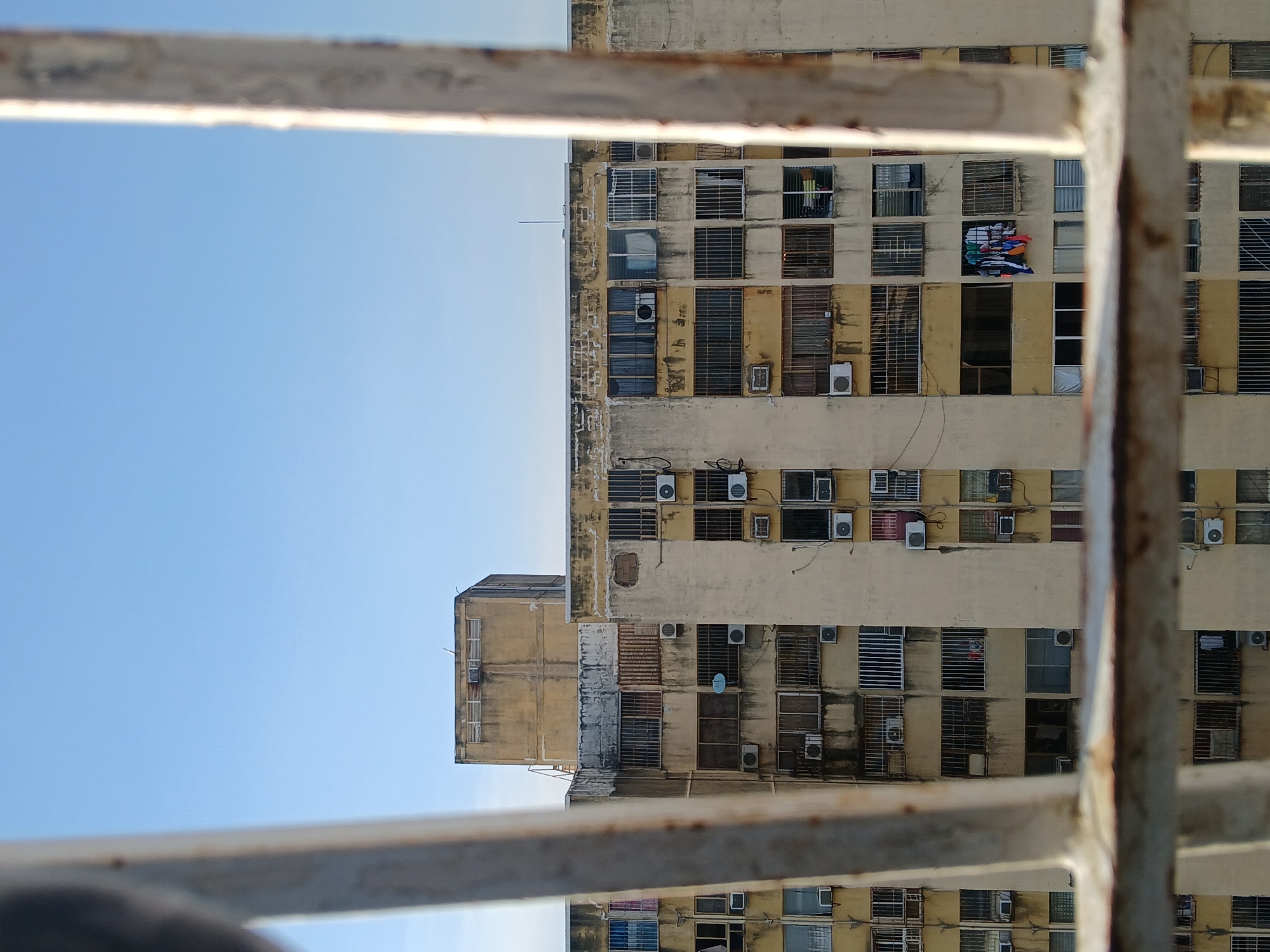 The image shows a high-rise building with multiple windows, viewed through what appears to be a metal railing. The building is off-white to yellowish, with many of the windows having metal bars. Several air conditioning units are attached to the building's facade. Clothes are hanging out of one of the windows. The sky is visible on the left of the frame. The building appears to be old and possibly in need of maintenance. Based on the prompt, the image depicts a scene in Ciudad Guayana, Venezuela.