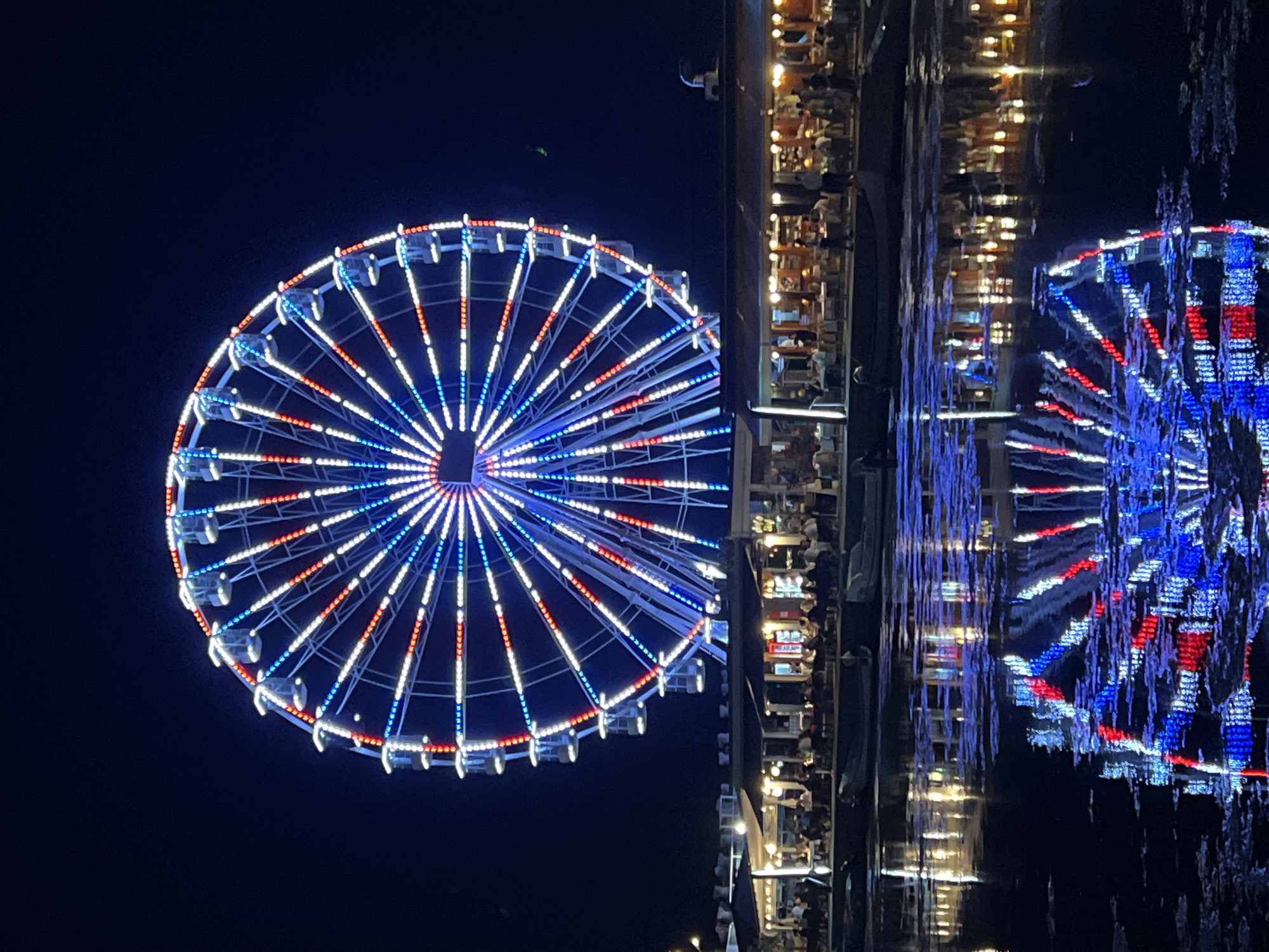 The image depicts a brightly lit Ferris wheel at night, reflecting in the calm waters below. The Ferris wheel is adorned with red, white, and blue lights, arranged in a radial pattern. Beneath the Ferris wheel, a waterfront promenade is visible, lined with buildings that appear to be restaurants or cafes, with illuminated tables and patrons seated outside. Boats are docked along the waterfront. The scene is set against a dark night sky. This Ferris wheel is a prominent landmark in the city of Constanța, Romania, located at the entrance of the Tăbăcărie Lake Park.