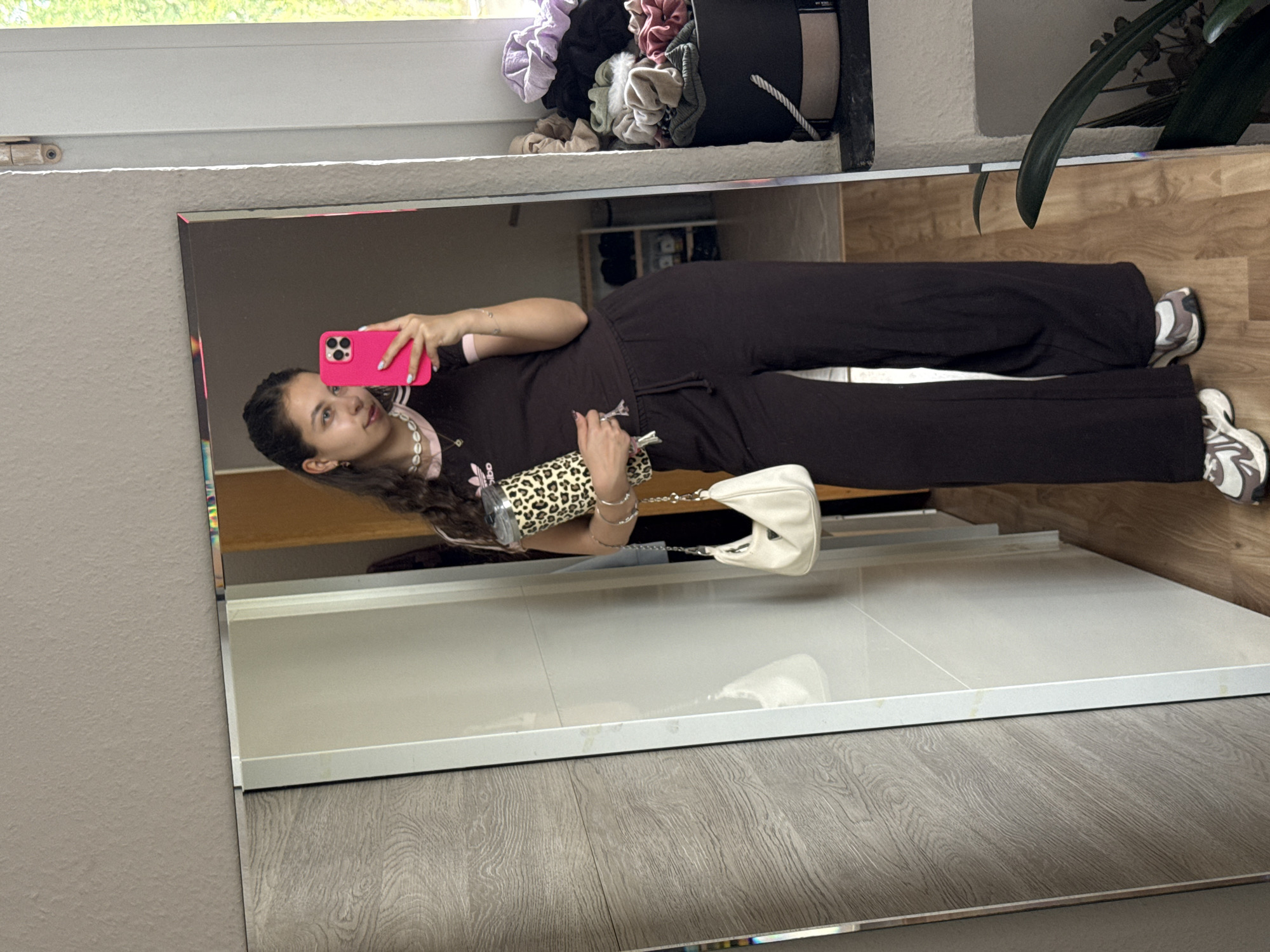 A young woman is taking a selfie in a mirror. She is wearing a brown Adidas tracksuit, a shell necklace, and white sneakers. She is holding a pink iPhone in her right hand and a leopard-print thermos and a white handbag in her left hand. The mirror is reflecting a white tiled shelf and a wooden floor. In the background, there is a bucket filled with hair scrunchies and a green plant. The photograph was taken in an unknown location in Bruchköbel, Germany.