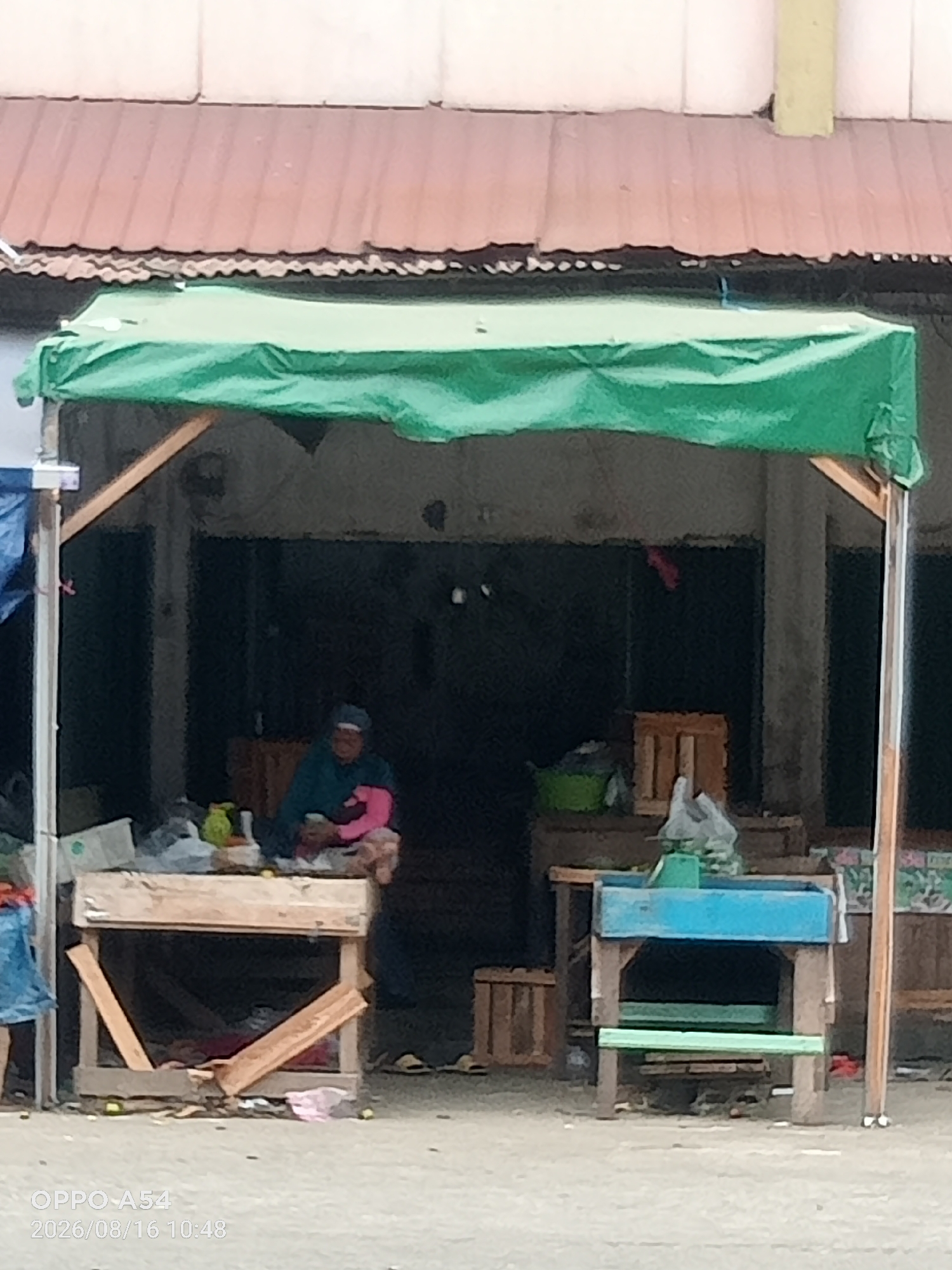 An outdoor market scene in Jambi, Indonesia, features a vendor and various goods. A person wearing a headscarf and a pink shirt is seated behind a wooden stall, appearing to be selling items. In front of the stall, there is a wooden stand with some fruits and other goods on display. To the right of the stall, a blue and wooden stand is visible, with a green bench underneath. The market area is covered by a green tarp supported by wooden poles, and a corrugated metal roof is seen overhead. The background appears to be a dark, open-air structure. The ground is paved with a light-colored material. The timestamp on the image indicates it was taken on August 16, 2026, at 10:48 AM.