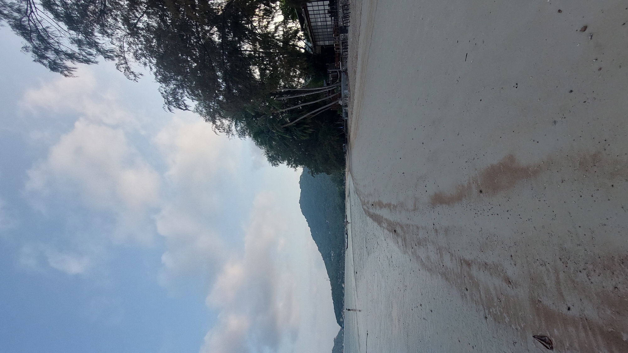 The image shows a beach scene in Koh Tao, Thailand. A sandy beach occupies the right half of the image, with darker sand indicating wetness near the water's edge. A line of trees and buildings sits along the beach. A mountain covered in vegetation rises in the background. The sky takes up the left side of the image, displaying a mix of blue sky and white clouds.
