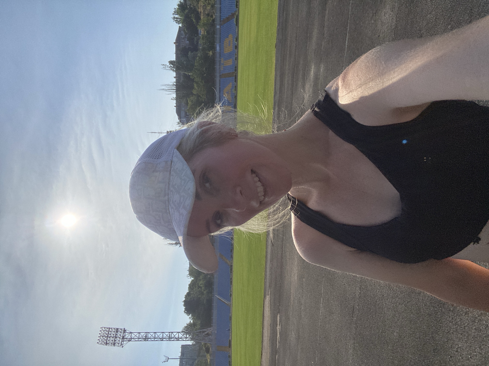 In an outdoor, daytime setting, a Caucasian woman with blonde hair and fair skin smiles at the camera. She is wearing a black tank top and a white baseball cap. The background features a stadium with blue and yellow seats, a green field, and a running track. A tall lighting tower stands to the left of the frame. The sun is visible in the sky.