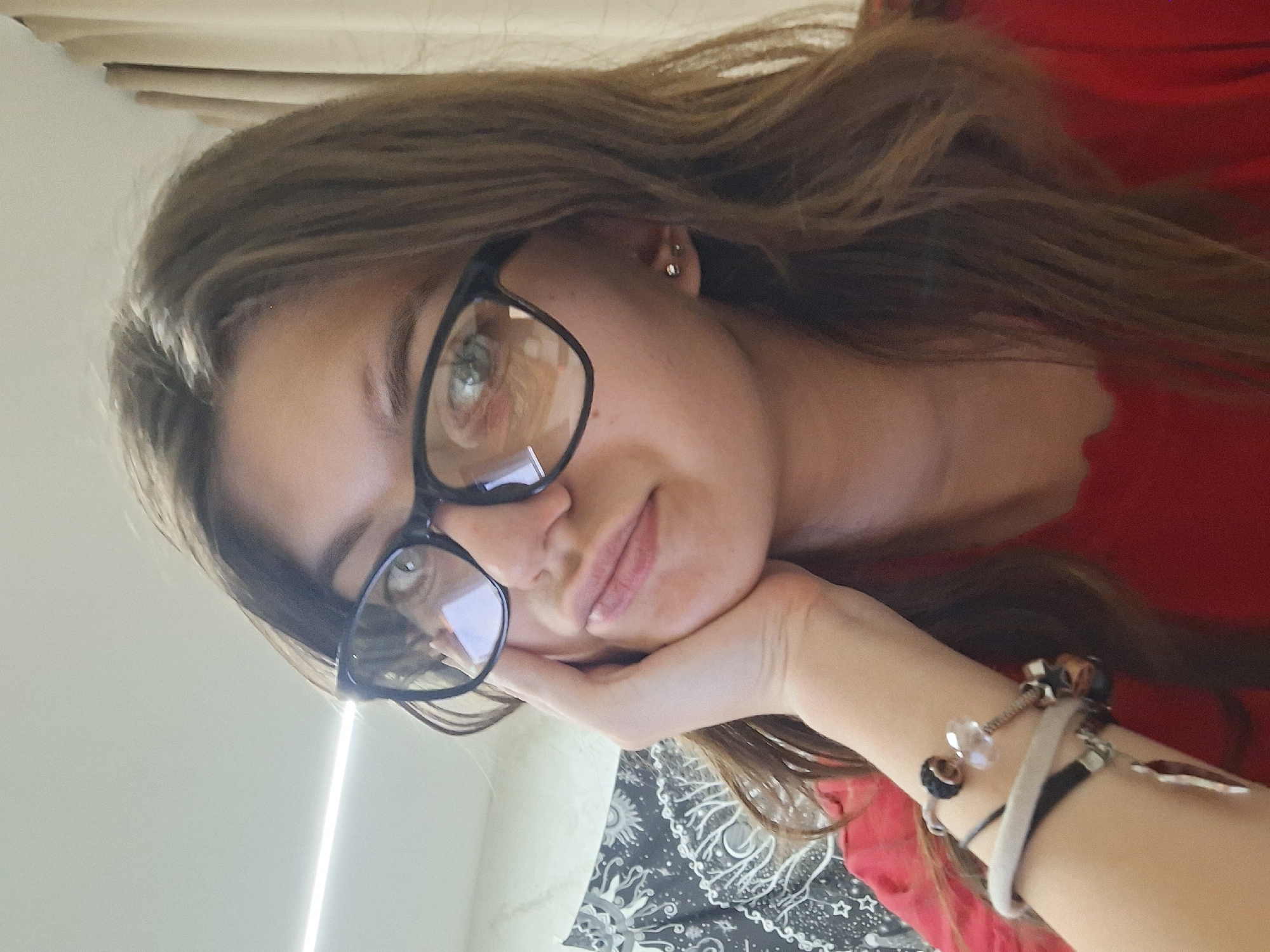 A woman with long brown hair and glasses is looking at the camera. She is wearing a red shirt and has several bracelets on her wrist. Her head is resting on her hand. A black and white patterned blanket is visible in the lower part of the frame. A light beam is visible on the left side of the image. The location is unknown, and there is no evidence to suggest it is Panagyurishte, Bulgaria.