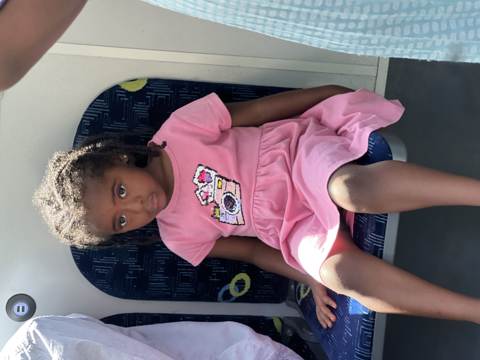A young Black girl with dark, curly hair is seated on a blue and white patterned seat, likely on public transportation such as a train or bus, given the visible USB charging port on the wall behind her. She is wearing a short-sleeved pink dress adorned with a sequined graphic of a camera. Her gaze is directed towards the camera with a serious expression. To her left, a person in a light-colored garment is partially visible, and to her right, another person in a light blue patterned garment is also partially in view. The lighting suggests it is daytime. The presence of the USB port and the seating arrangement indicate an urban transportation environment. While the specific city is not identifiable from visual cues alone, the prompt specifies Kiryat Bialik, Israel.