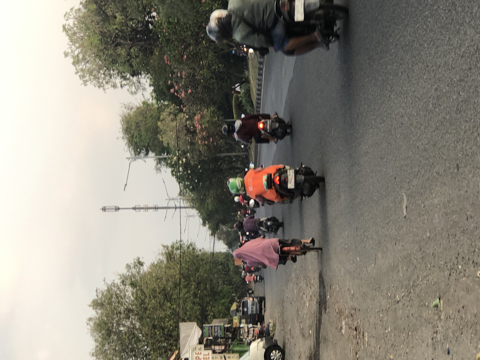 The image depicts a street scene in Gelam, Indonesia, characterized by a moderate flow of traffic, predominantly motorbikes. A lone cyclist, a woman wearing a pink hijab, is prominent in the foreground, riding a bicycle in the same direction as the motorbikes. Several motorbikes are visible, with riders and passengers wearing helmets, some in brightly colored jackets. One rider wears an orange jacket and a green helmet, suggesting a delivery service. Buildings with signage indicating shops line the left side of the road, with parked vehicles and a white van. The road surface is asphalt, with some debris scattered on the shoulder. Trees with green foliage and some flowering bushes line the right side of the road, creating a somewhat urban-rural landscape. Overhead, a cell tower and streetlights are visible against a hazy sky, indicating it is either early morning or late afternoon. The overall impression is of a typical day in a bustling Indonesian town.