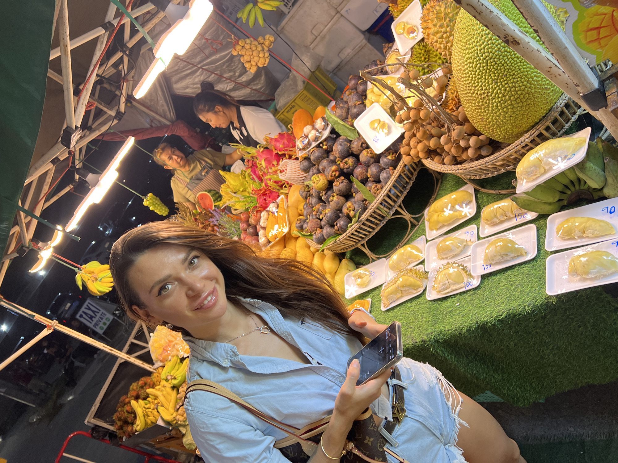 A woman with long brown hair, wearing a light blue button-down shirt and ripped denim shorts, holds a smartphone in her right hand in an image taken at a fruit stand in Baan Bo Phut, Thailand. She smiles at the camera. The fruit stand is overflowing with tropical fruits, including durian, jackfruit, mangosteens, dragon fruit, mangoes, bananas, and longans. Two individuals, presumably vendors, are working in the background. Fluorescent lights illuminate the stand, while a 'TAXI PRIVATE' sign can be seen in the background. The fruit is arranged in baskets and on display, with prices marked on some of the durian.