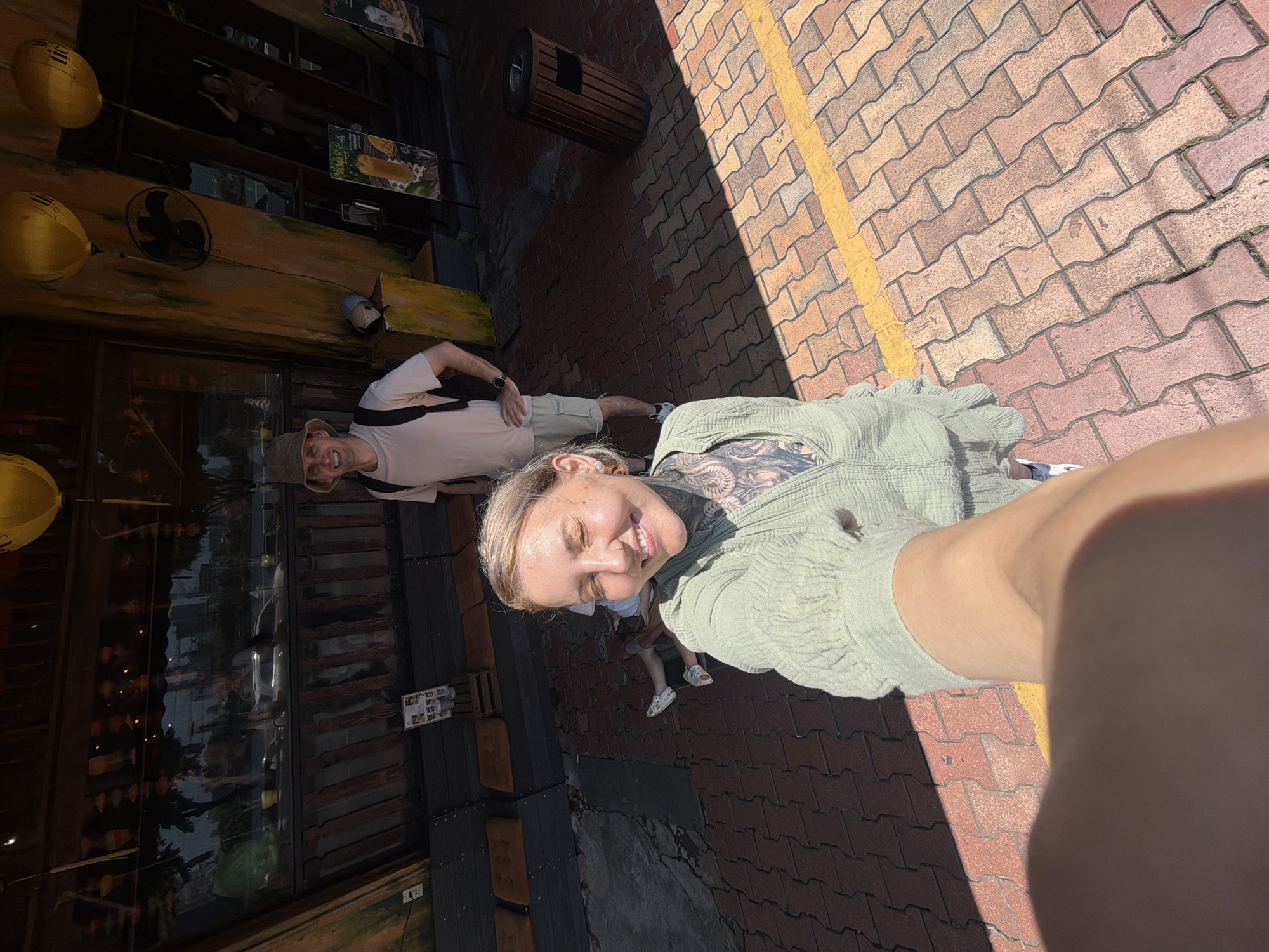 The image captures a woman in a green shirt taking a selfie outdoors in Đà Nẵng, Vietnam. She is smiling at the camera. Behind her stands a man in a khaki hat and white t-shirt with a shoulder bag. A child in sandals is also present near the woman. They stand on a brick sidewalk with a yellow line. Behind them, on the left, is a building with dark, reflective glass windows. Lanterns hang above the entrance of the building. A waste container sits against the building and sidewalk.