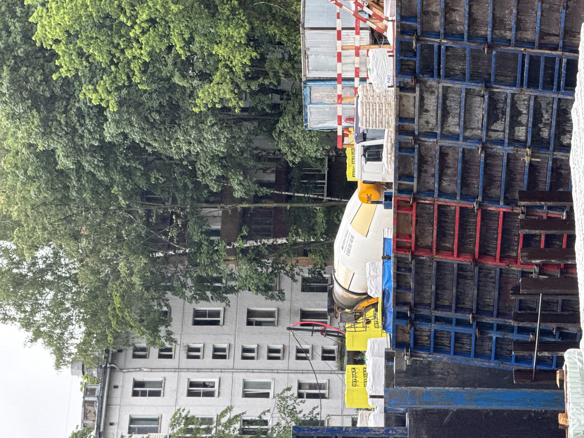 This image shows a construction site in Moscow, Russia, with a concrete mixer truck in the foreground. The truck is parked next to a large, blue and red metal formwork structure, which is likely being used for concrete pouring. In the background, there are several apartment buildings and trees. The sky is overcast, suggesting it might be a cloudy day. There are stacks of white bags, possibly cement, near the concrete mixer. The overall scene is typical of urban construction work.