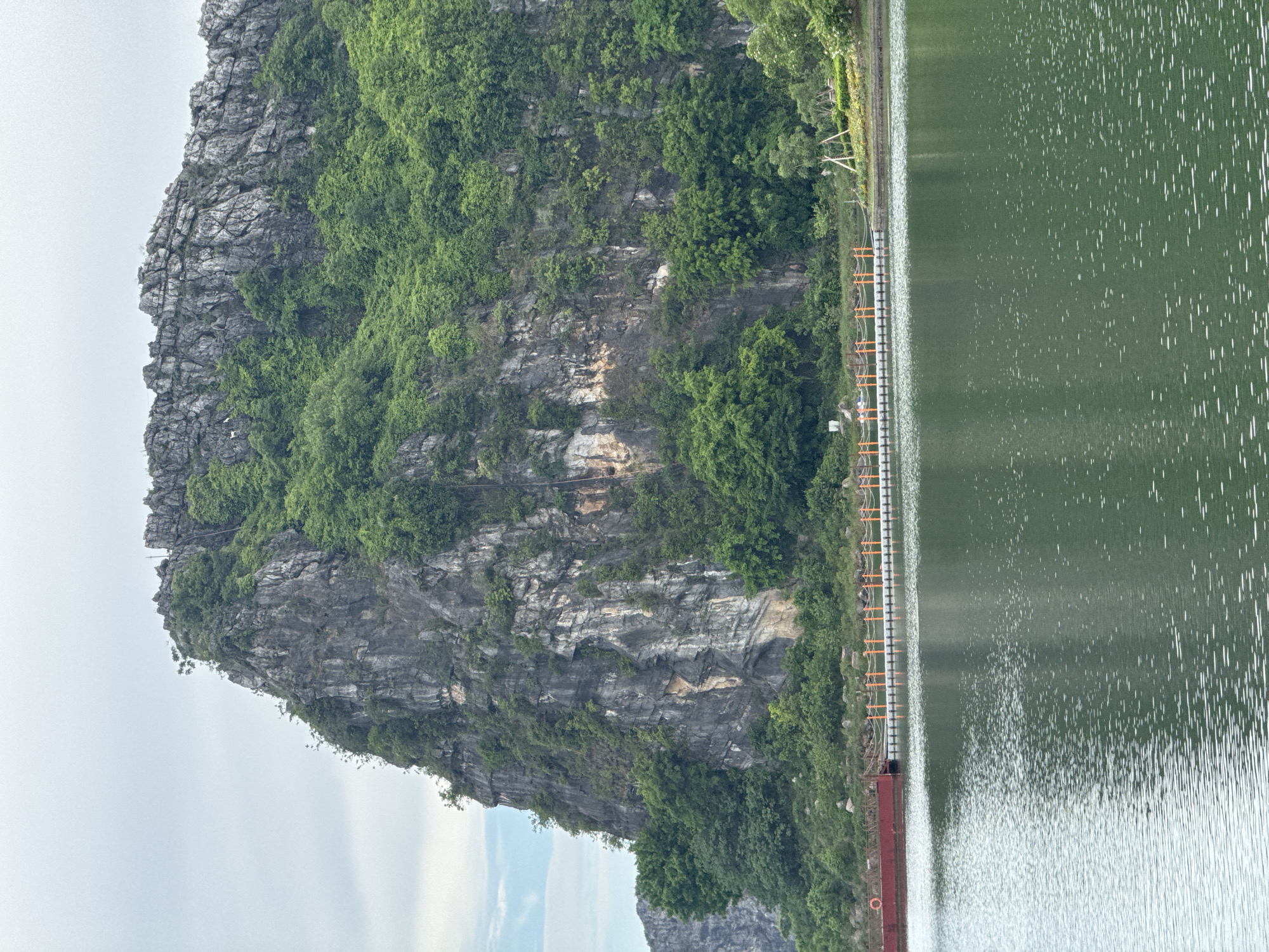 An overcast day frames a steep, rocky karst mountain covered in lush green vegetation. The mountain dominates the upper half of the image. In the foreground, a calm, green body of water reflects the sky and the verdant hillside. Along the edge of the water, a man-made walkway extends across the scene. This walkway is supported by a series of white floats and is lined with orange railings. A red barge is docked to the left of the walkway. Two people are visible on the walkway, appearing to be walking. The location is Hoa Lư Ward, Vietnam, a region known for its dramatic karst landscapes and waterways.