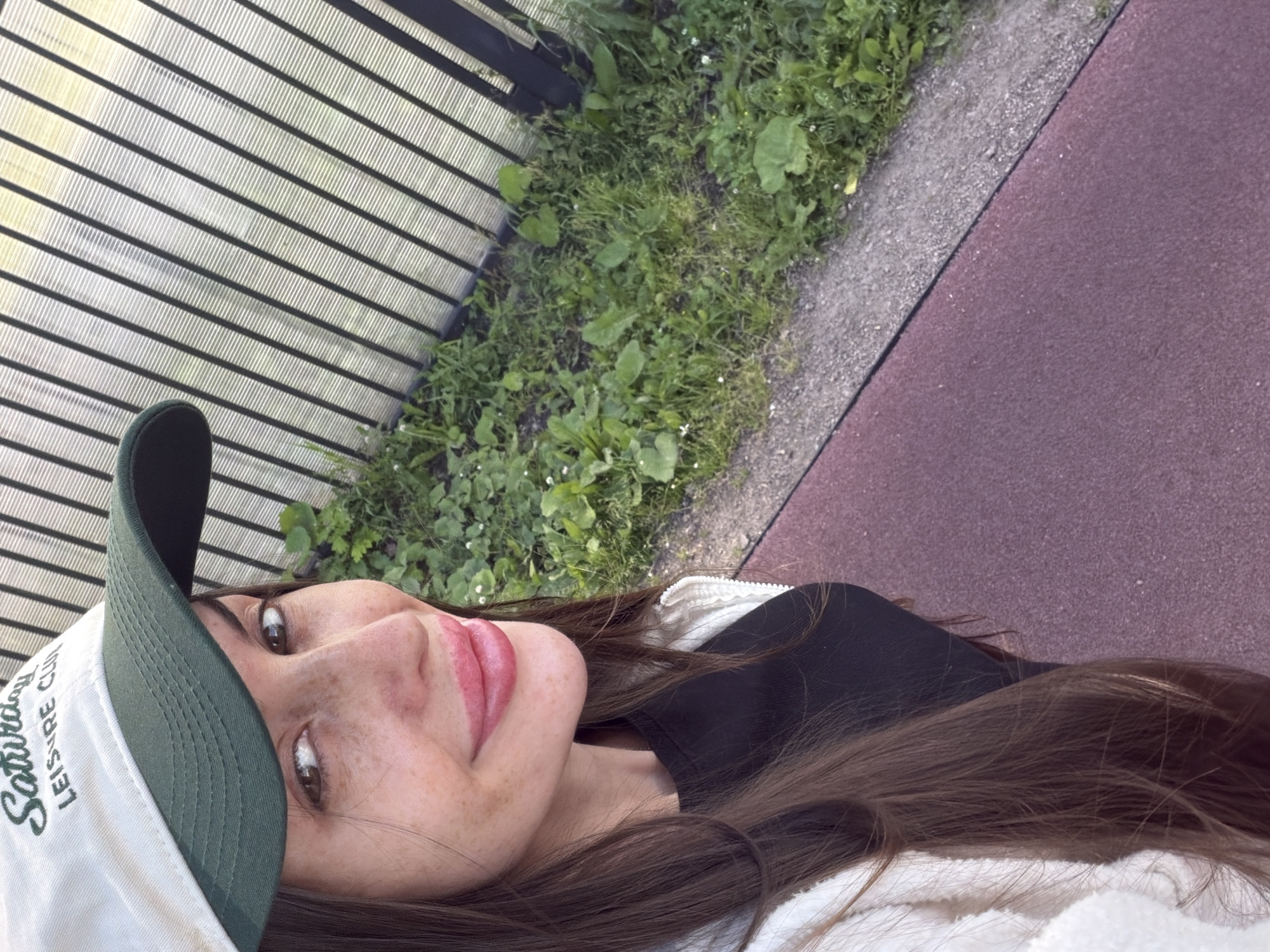 The image shows a close-up, sideways selfie of a young woman with long, dark brown hair, freckles, and light brown eyes. She is wearing a white baseball cap with green lettering that reads "Saturday Leisure Club" and a black t-shirt. Her lips are slightly parted and appear to be painted with a pink lip gloss. The lighting suggests it is daytime, and the scene is outdoors, with elements of nature visible. In the background, there is lush green grass with some small white flowers, and beyond that, a reddish-purple path. To the left, there is a slatted fence or wall, possibly made of wood or plastic. The overall impression is that of a casual, relaxed moment. The text on the cap is clearly legible. The provided location context of Saint Petersburg, Russia, does not offer specific visual cues within the image itself to confirm or deny this. There are no other people or objects of significance in the frame. The woman's expression is neutral, with a slight upward glance, contributing to the relaxed, candid feel of the photo.