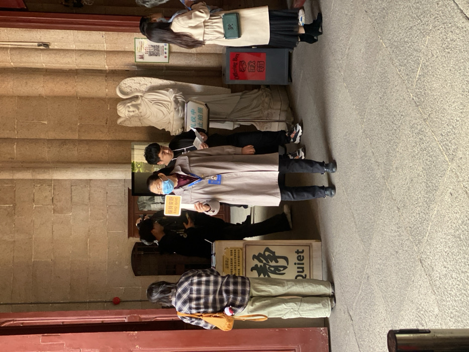 This image, taken in Guangzhou, China, depicts an indoor or covered outdoor area of a building, likely a temple, church, or historic site. Several people are present, appearing to be queuing or gathered respectfully.
In the foreground, a person with long dark hair, wearing a plaid shirt and light pants, stands with their back to the camera, carrying a light brown bag. Adjacent to them, a person in a black jacket is visible. The central figure is an attendant, dressed in a light trench coat, dark pants, and a blue face mask, holding a yellow sign that reads "保持安静" (Keep quiet) and "Keep quiet." Behind this attendant, another individual in dark clothing is partially visible. Further back, a young woman with long dark hair, wearing a light beige coat and black pleated skirt, stands near a grey "Offering Box" (奉献箱).
The setting features sturdy, light brown stone walls and a paved floor. A white angelic statue with wings is prominent behind the attendant. Next to the statue, a white sign states "礼仪中禁止拍照" (No photography during ceremony). On the ground, near the masked attendant, a wooden sign prominently displays the Chinese character "静" (Jìng - quiet/calm) and the English word "Quiet." The presence of an offering box and signs requesting silence and prohibiting photography during ceremonies suggests a place of worship or a site requiring solemnity. The activity appears to be an orderly visit or attendance at a quiet event. The scene is illuminated by natural, albeit somewhat dim, daylight.